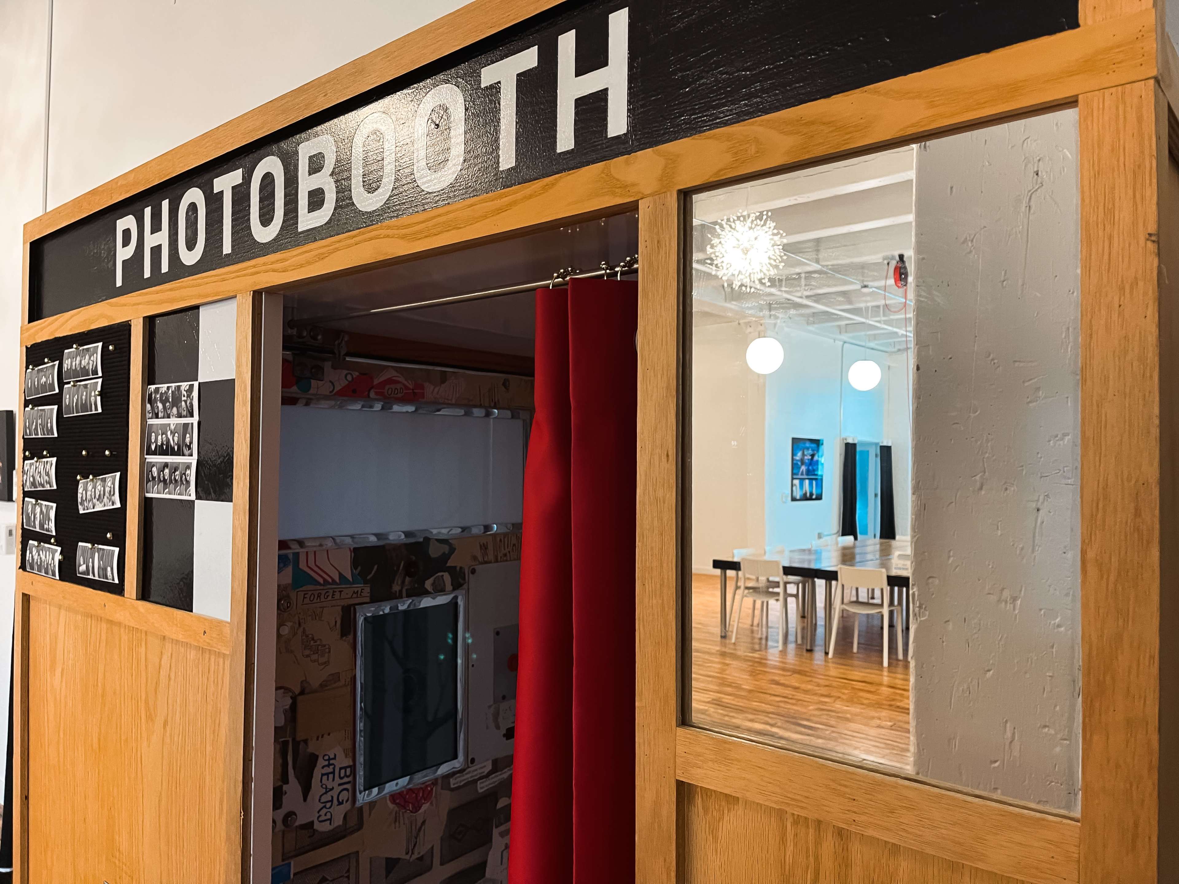 The image shows a wooden photobooth with a red curtain and a series of black-and-white photos displayed on its side, set in a bright interior space with tables and chairs visible in the background.