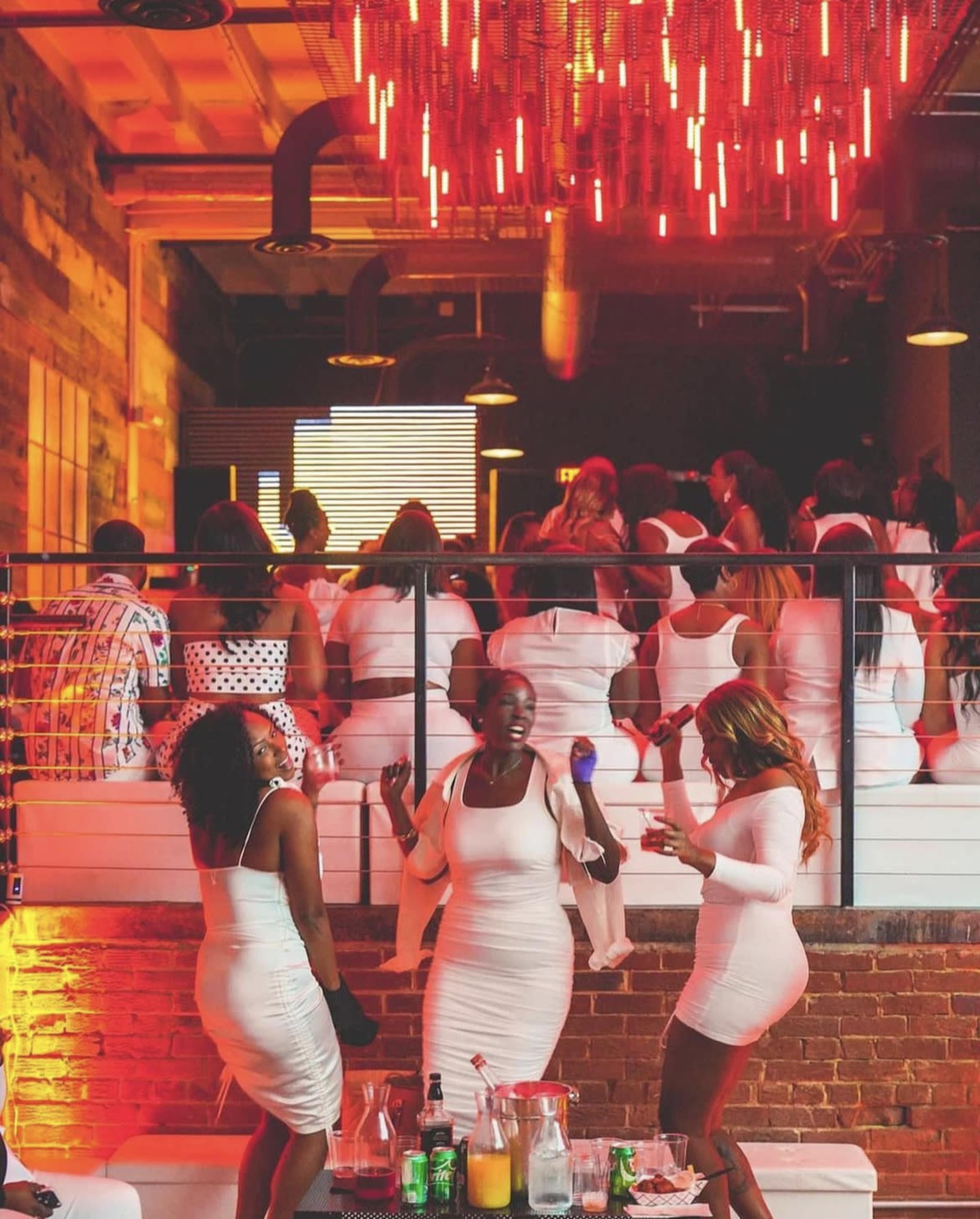 A group of people wearing white dresses socialize and dance in a vibrant venue with red lighting and a large audience in the background.