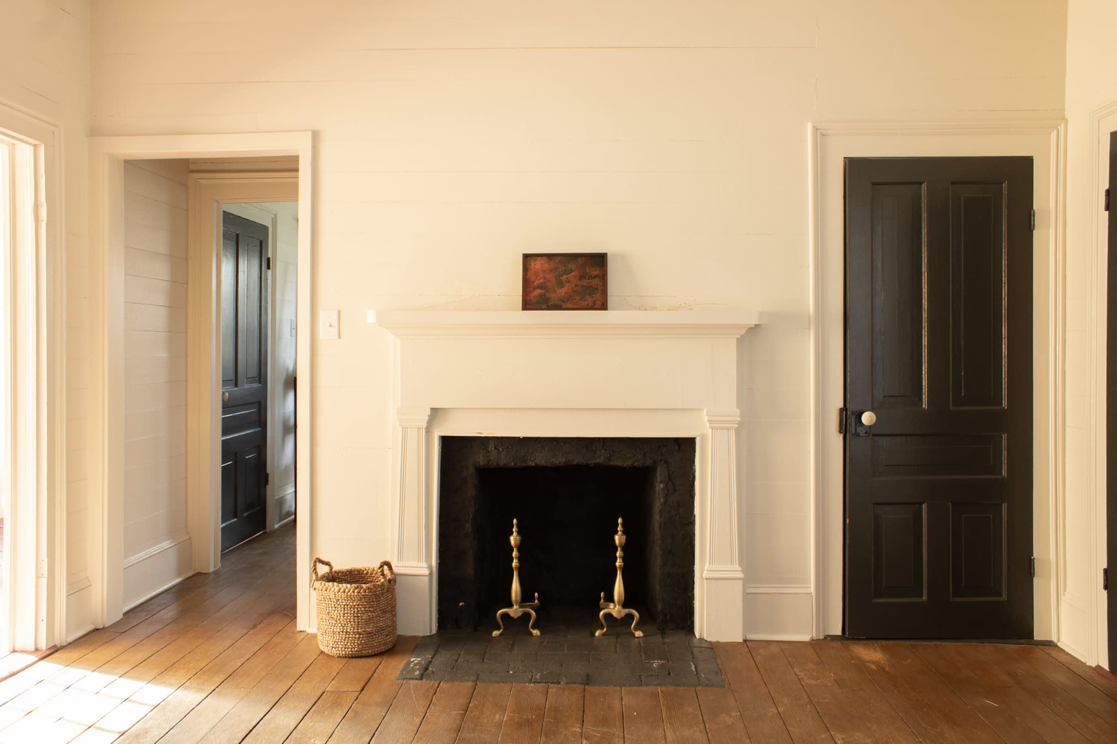 A simple room features a white-paneled wall with a fireplace, black doors, and a woven basket on the wooden floor.