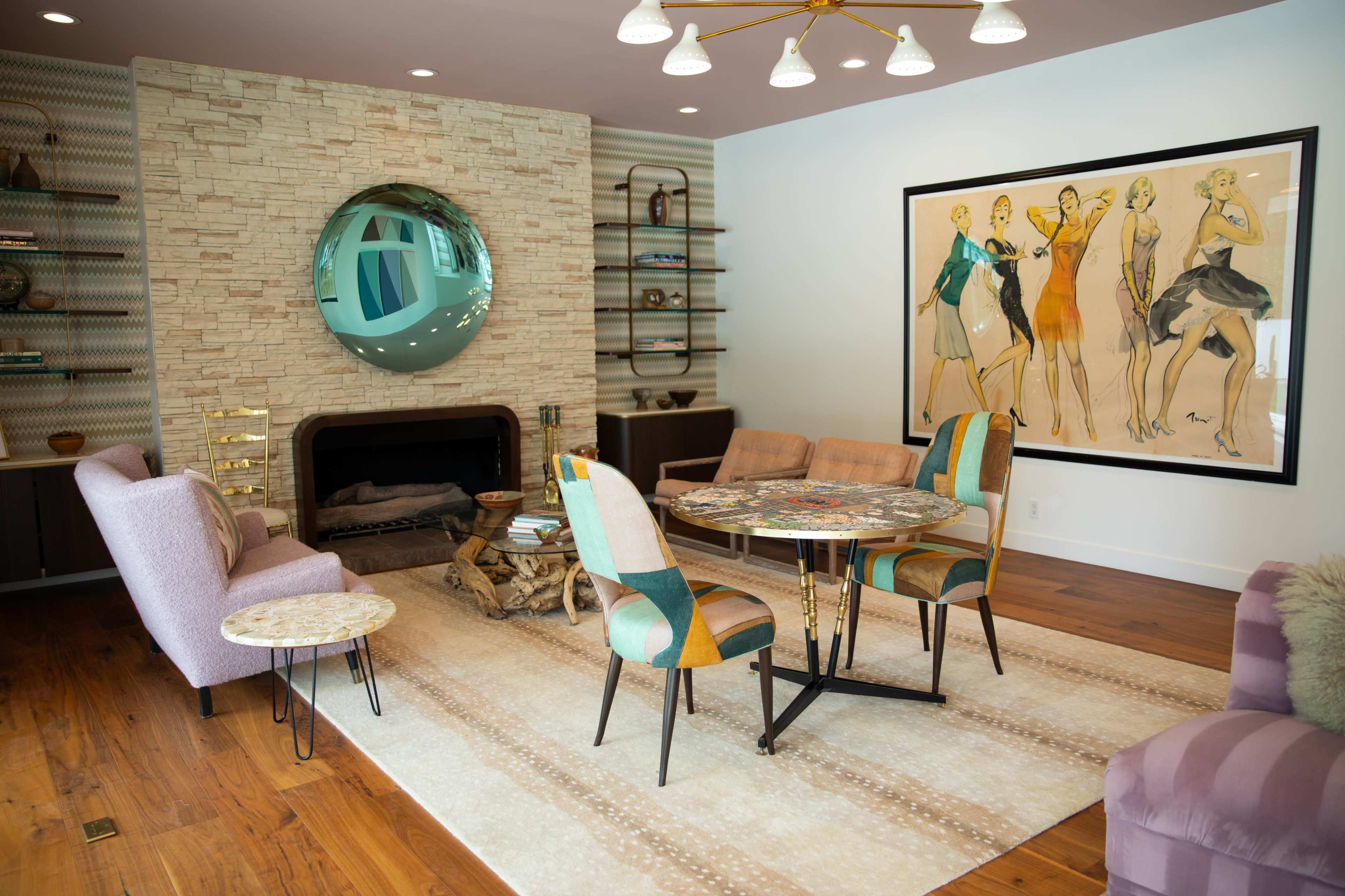 The image shows a modern living room with a stone feature wall, a circular mirror, a round dining table with colorful chairs, and a framed artwork of fashion models.