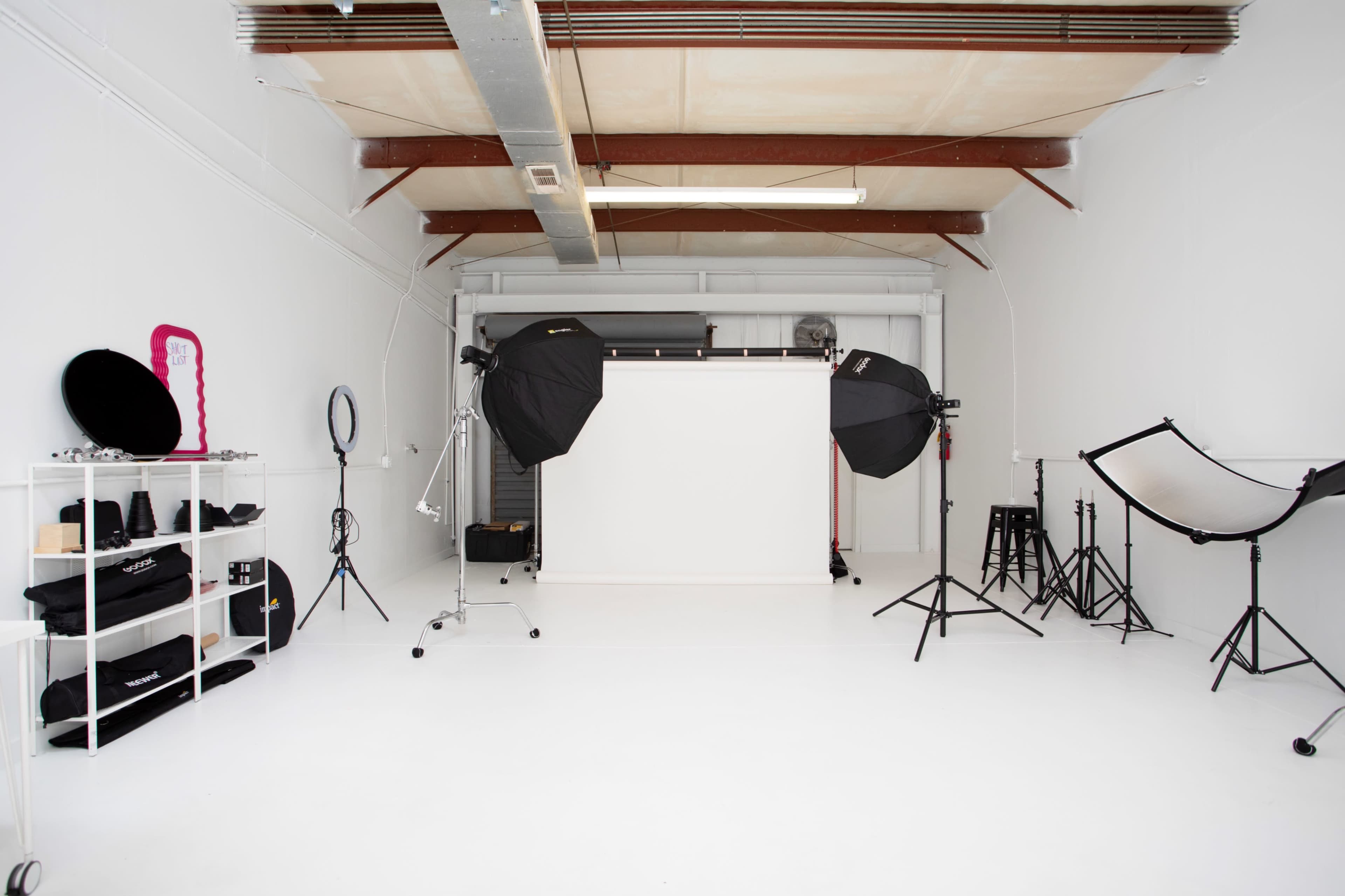 The image shows a photography studio with a white backdrop, lighting equipment, and various accessories organized on shelves.