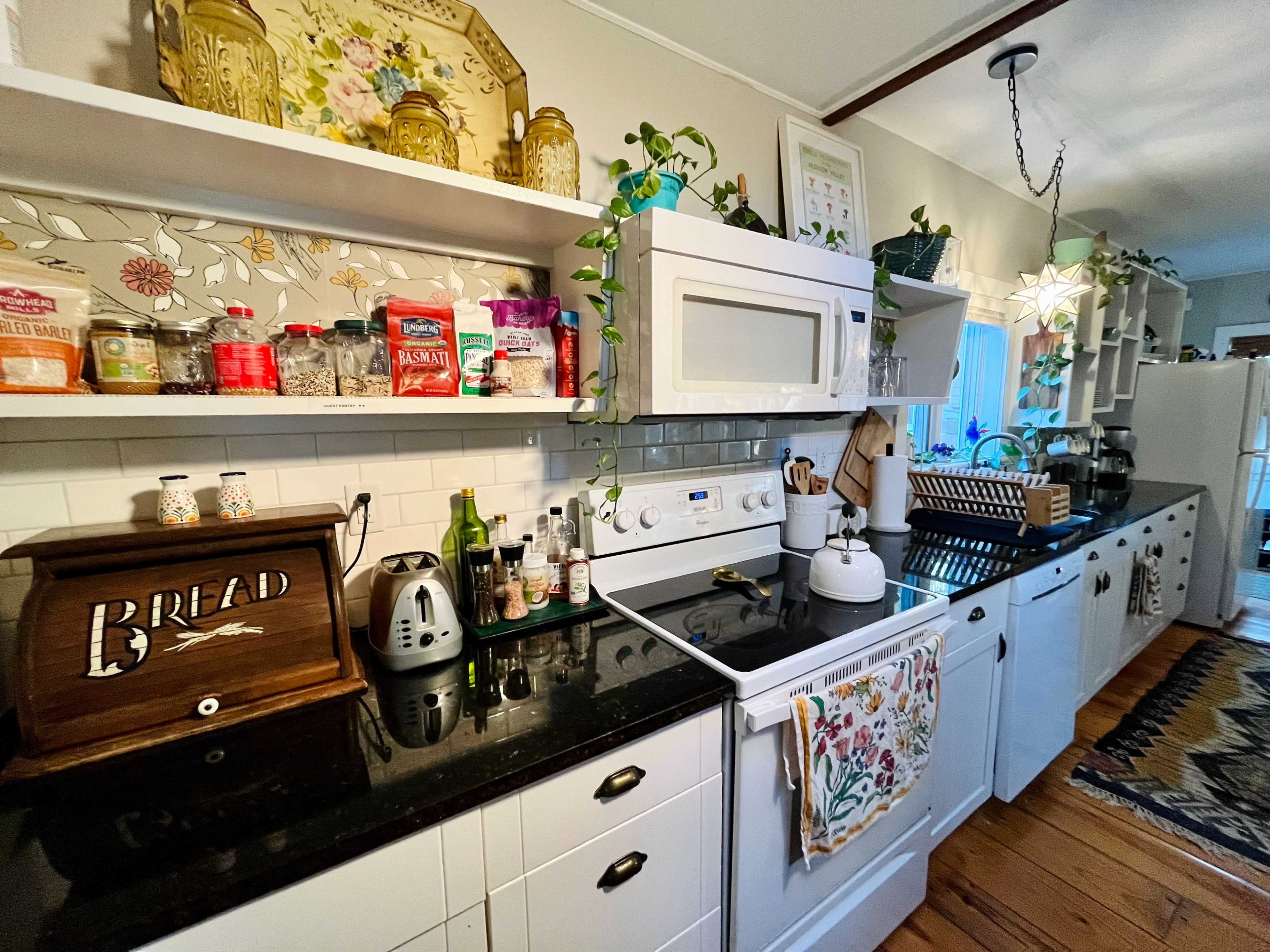 A kitchen features a black countertop with a stove and microwave, surrounded by white cabinets and shelves displaying jars and decorative items, along with a coffee maker and a bread box.