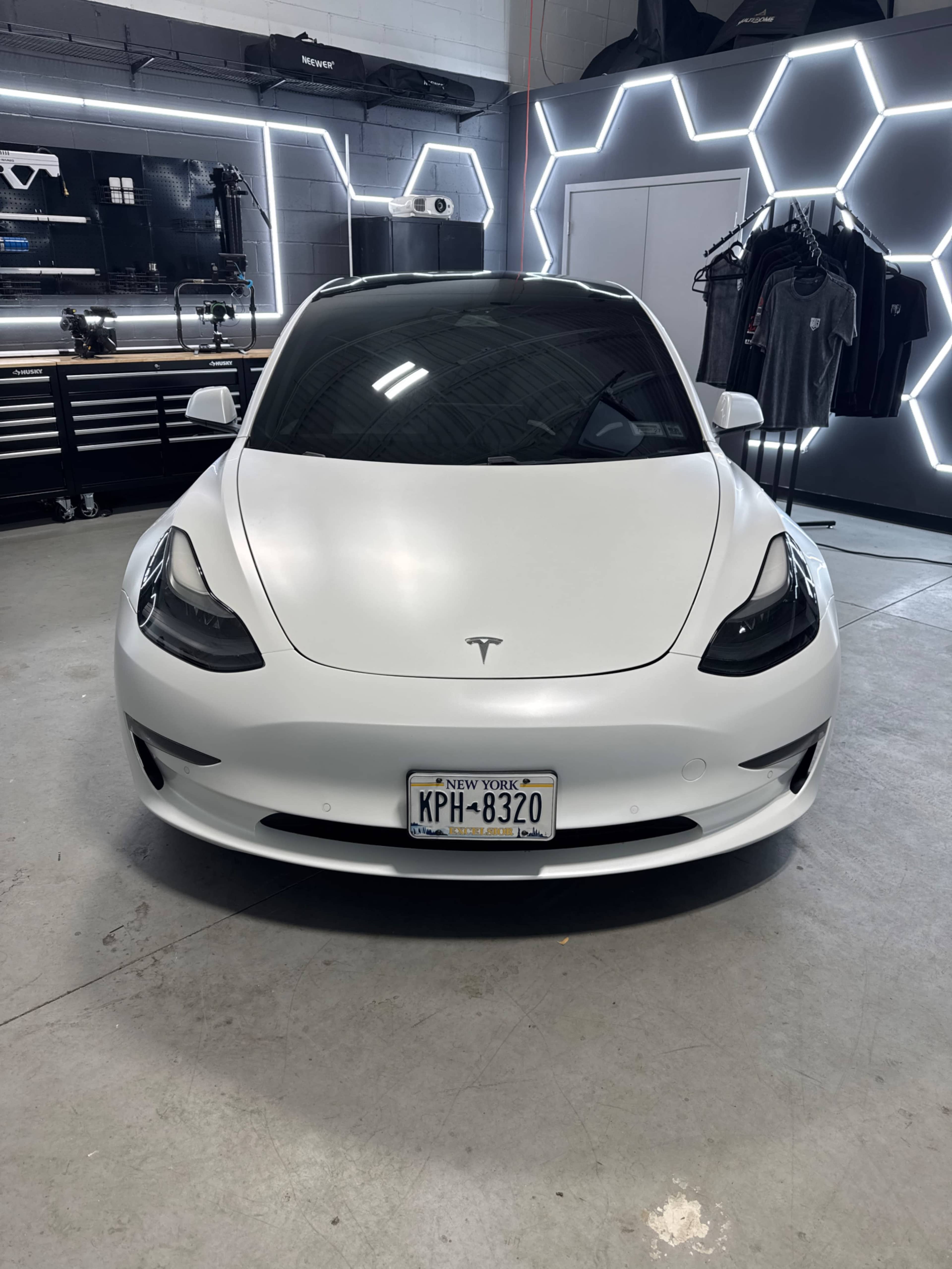 A white Tesla Model 3 is parked in a garage with a modern backdrop featuring geometric lighting and apparel displayed on racks.