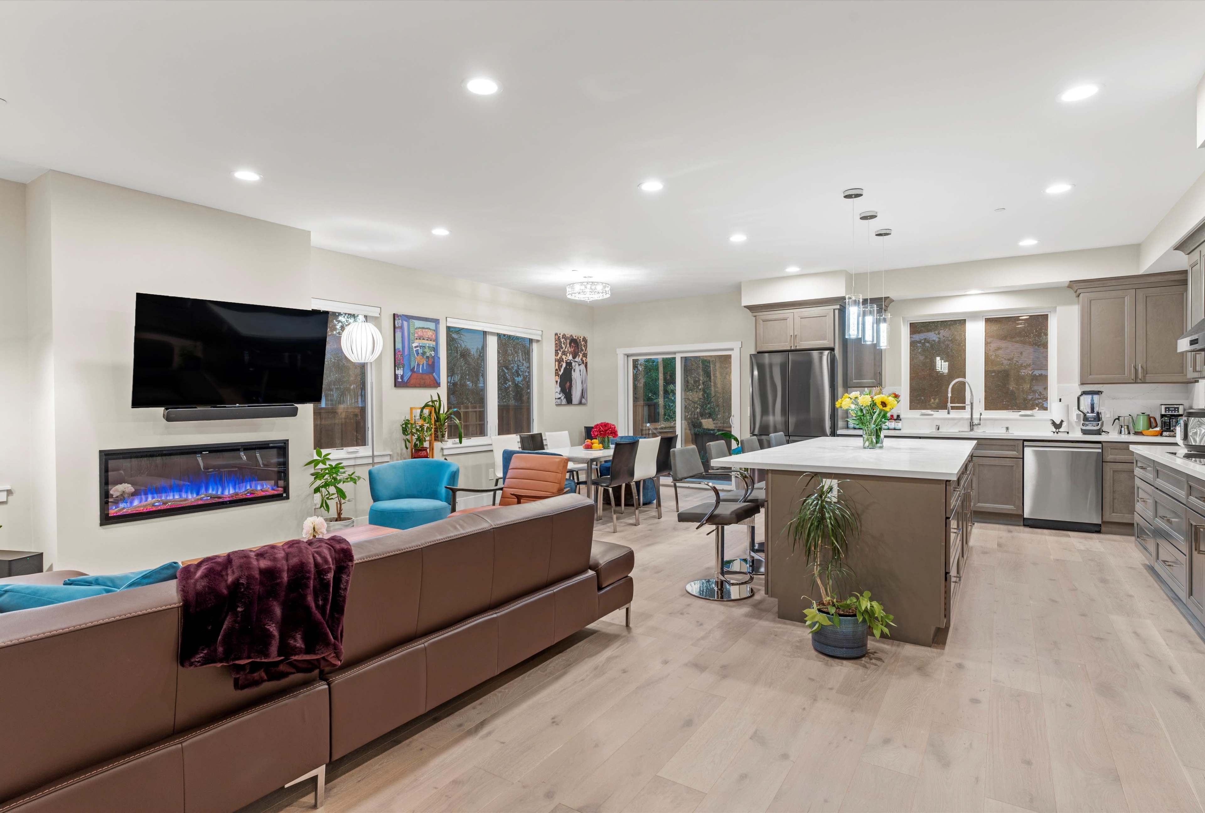 A modern open-concept living space features a kitchen, dining area, and seating lounge with large windows and a mounted television.