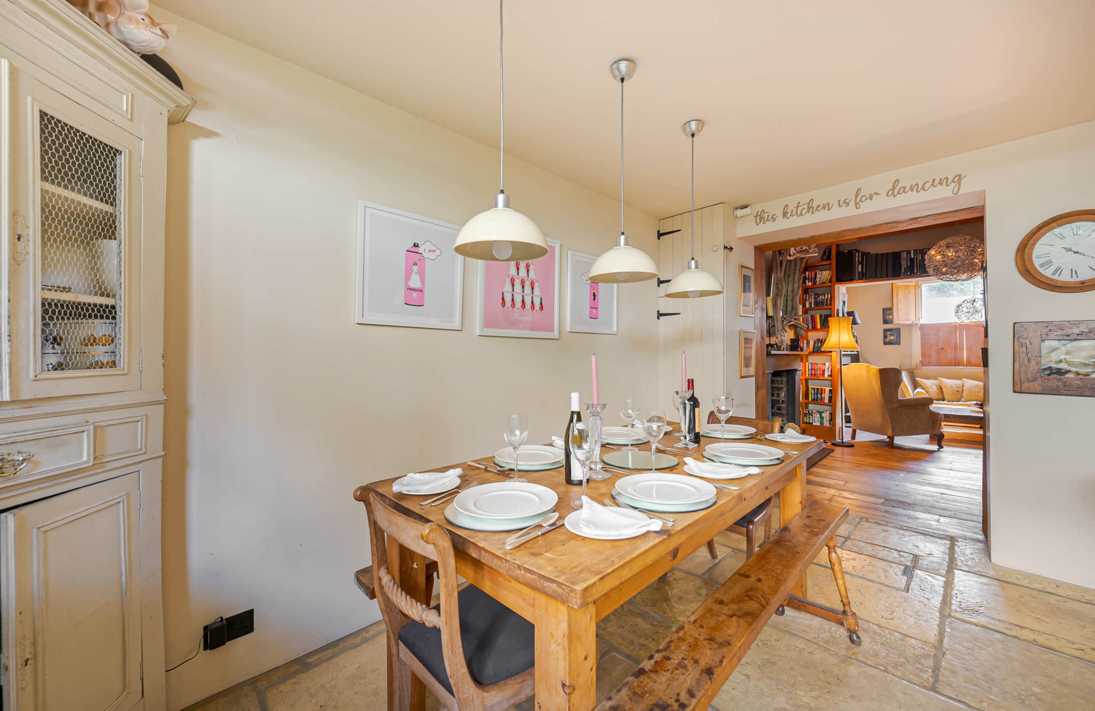 A wooden dining table is set with plates and glassware in a cozy kitchen area with light-colored walls and a doorway leading to a living space.