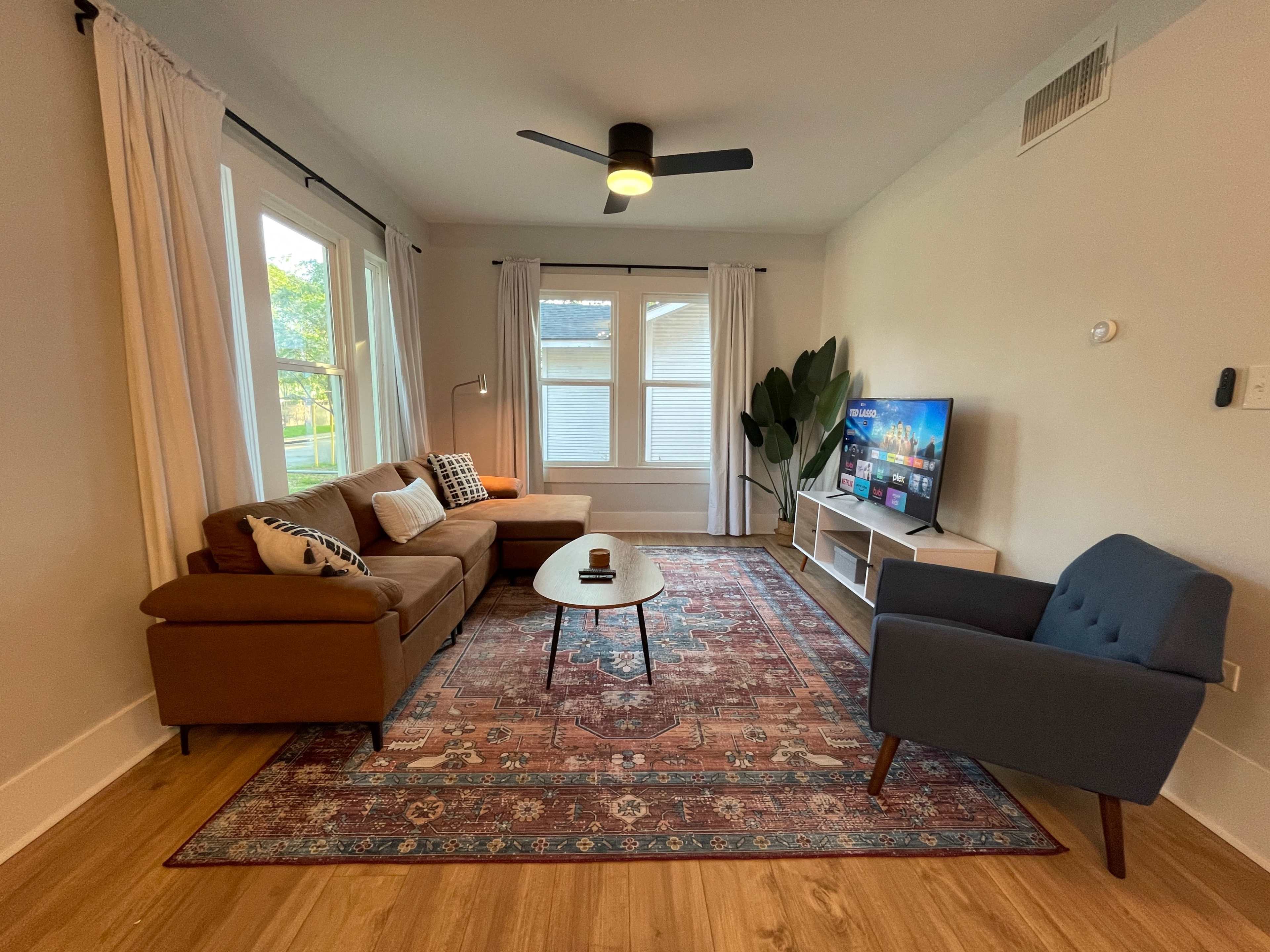 The living room features a brown sofa, a blue armchair, a round coffee table, and a television, all arranged around a patterned rug under a ceiling fan and natural light from two windows.