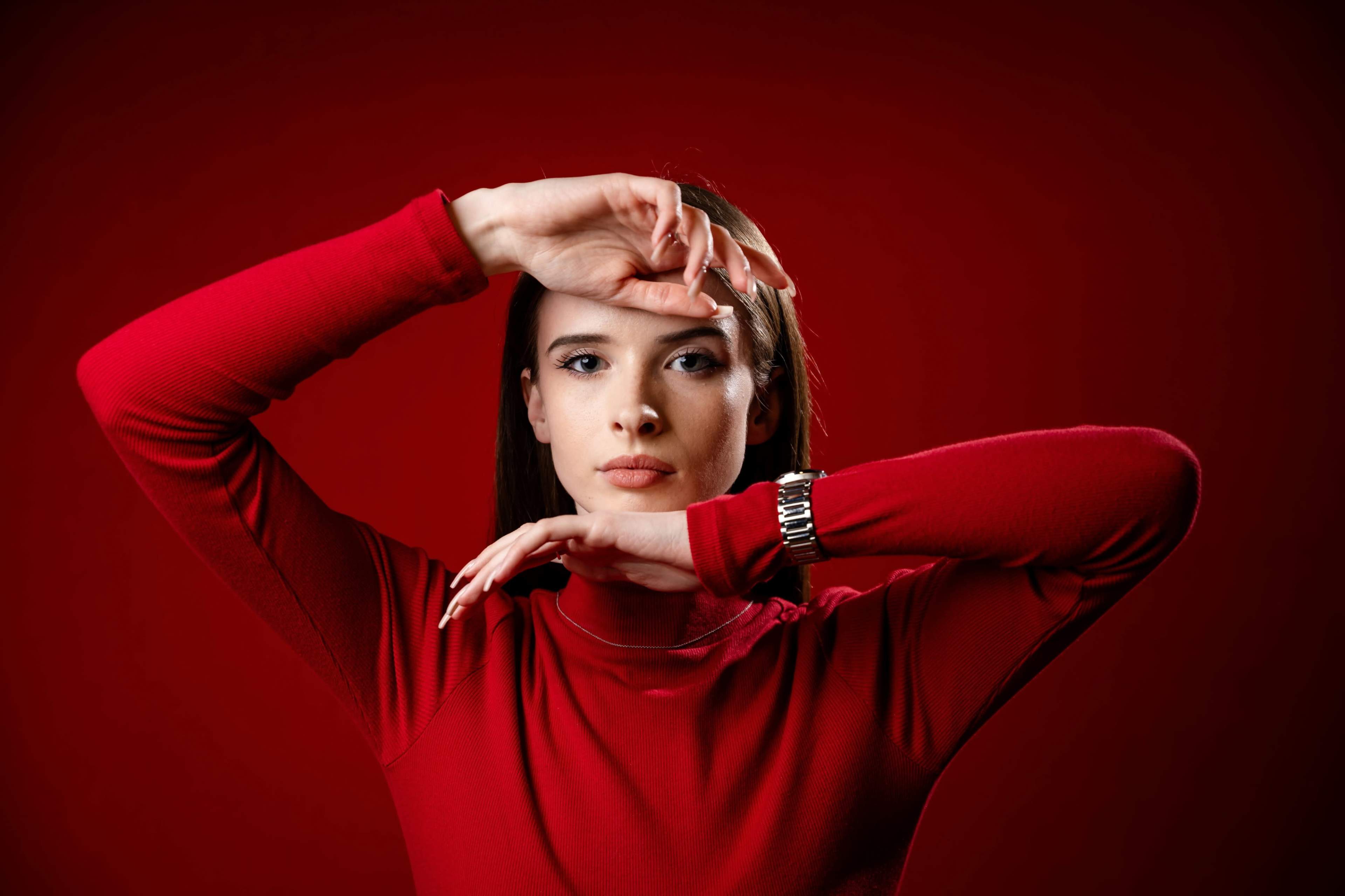 A woman in a red long-sleeve top poses with her arms raised in front of a solid red background.