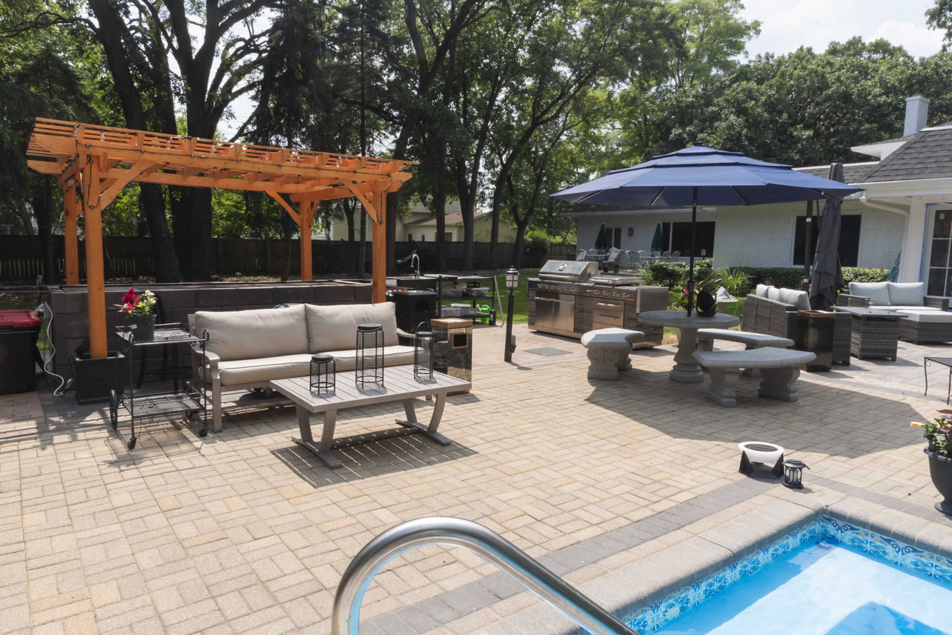 The scene shows a backyard patio with a pool, outdoor furniture, a grill area, and a pergola set amidst trees.