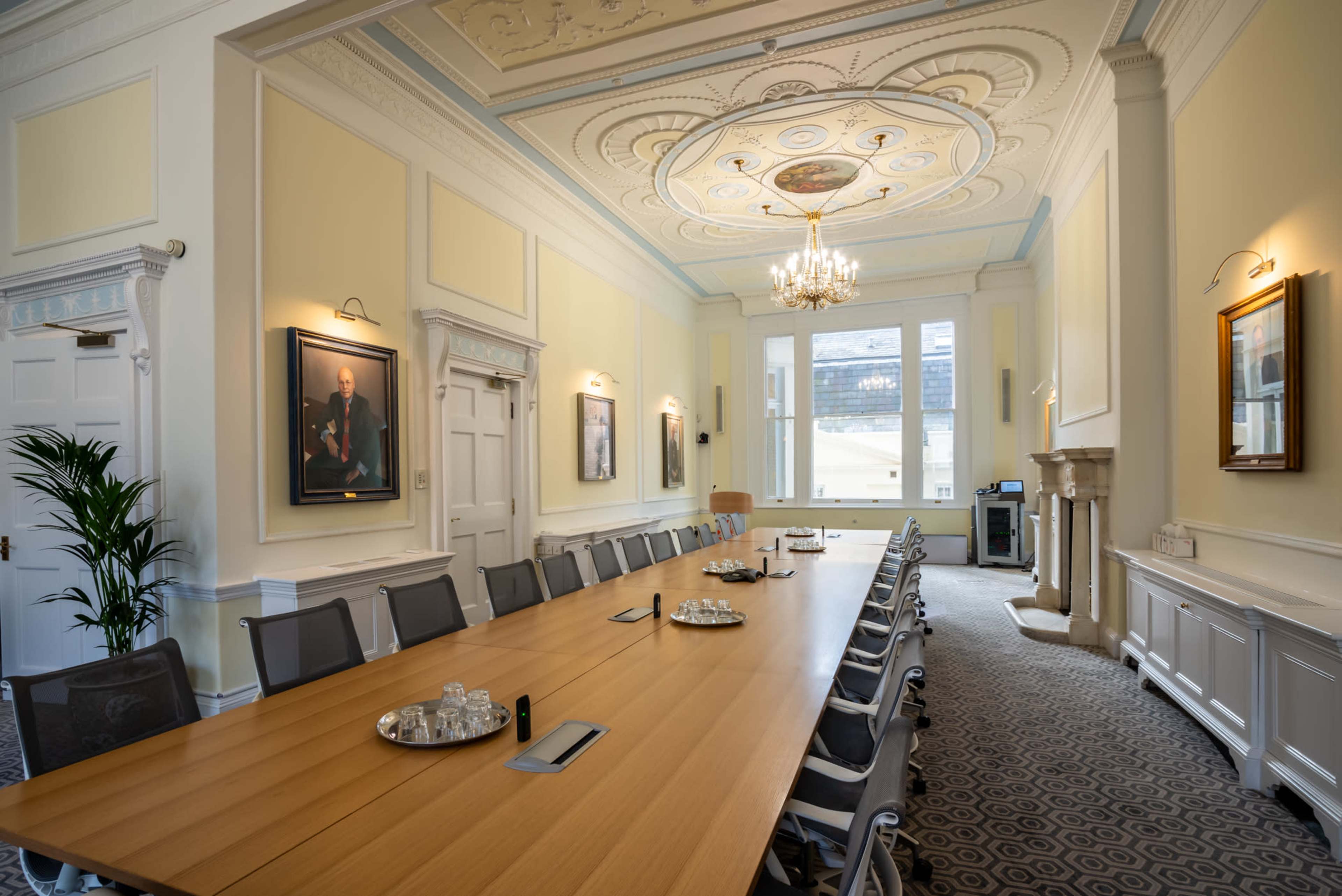 A spacious conference room is illuminated by a chandelier, featuring a long wooden table surrounded by chairs and adorned with framed portraits and large windows.