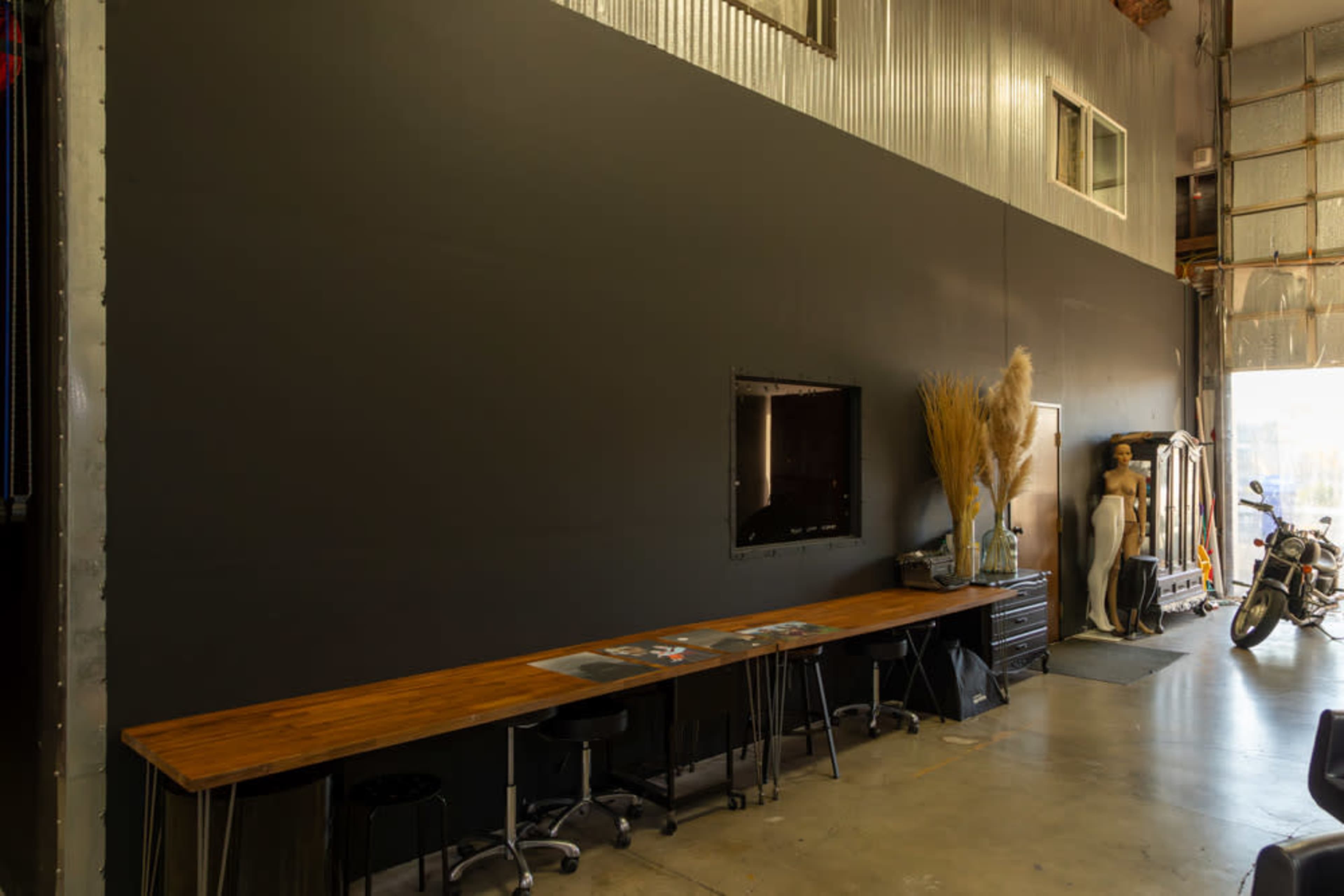 The image shows a spacious industrial interior with a long wooden table, a black wall, a television mounted at the center, and various items including a mannequin and a motorcycle.