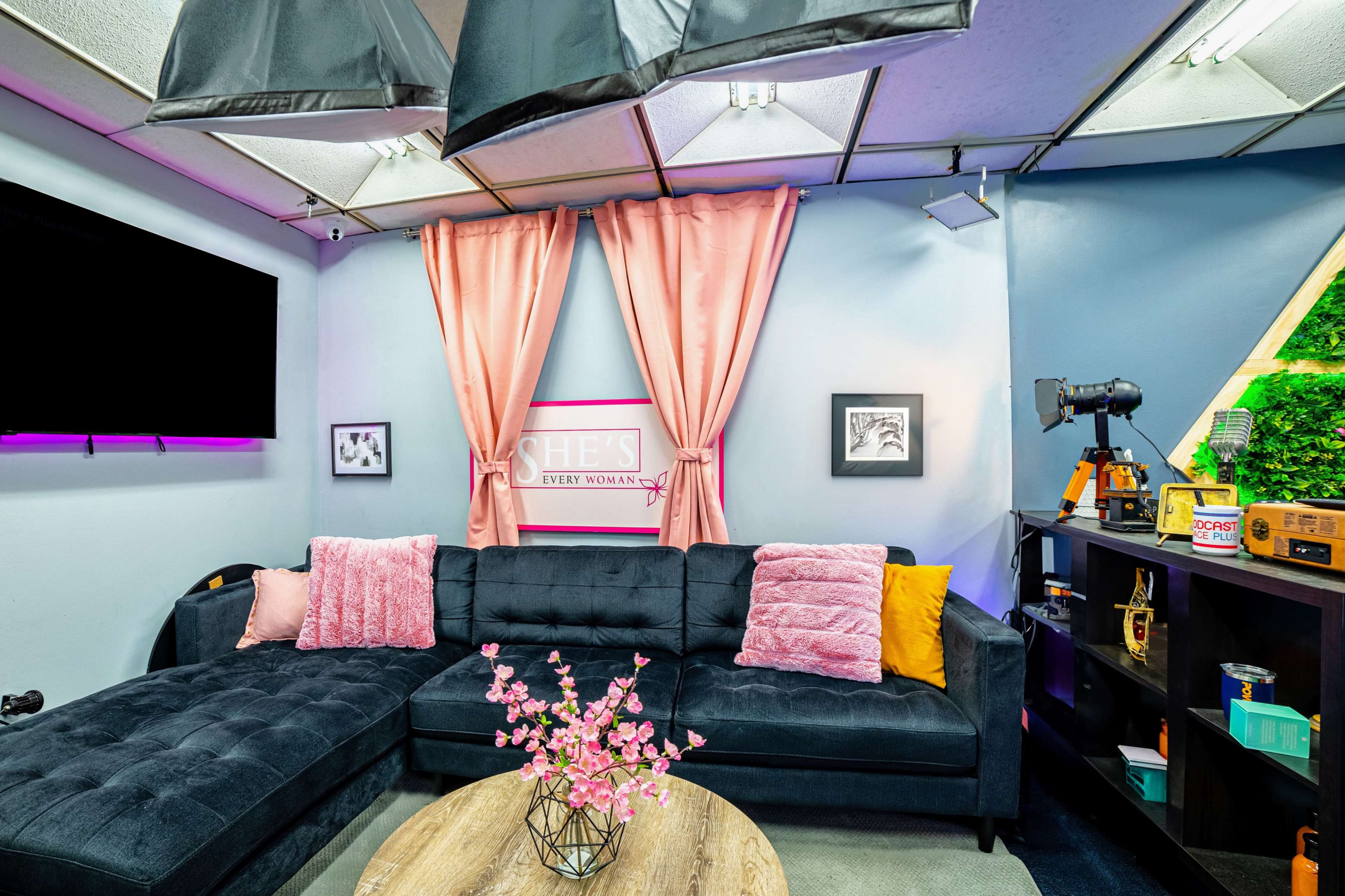 The image shows a modern living space with a dark sectional sofa, decorative pillows, a round wooden coffee table, and a backdrop featuring pink curtains and a framed sign.
