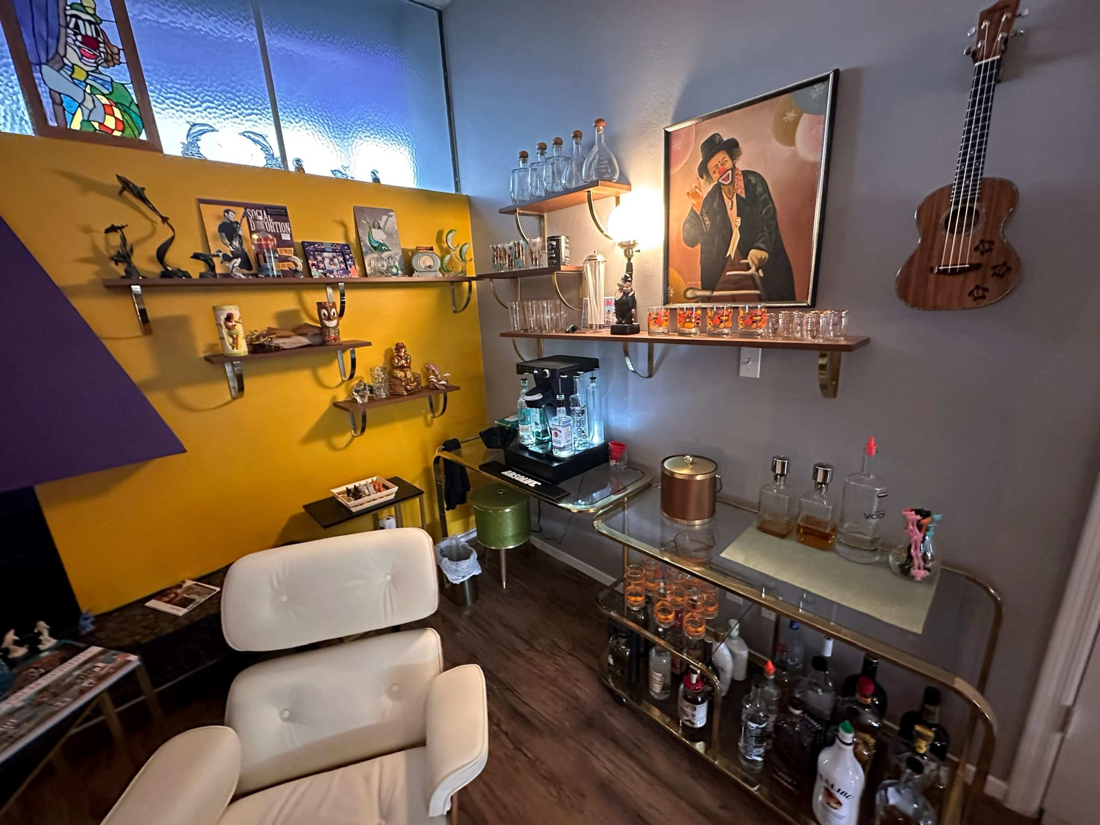 A stylish room with a yellow wall featuring shelves holding decorative items and a drink station equipped with bottles and glassware, alongside a vintage chair and a ukulele on the wall.