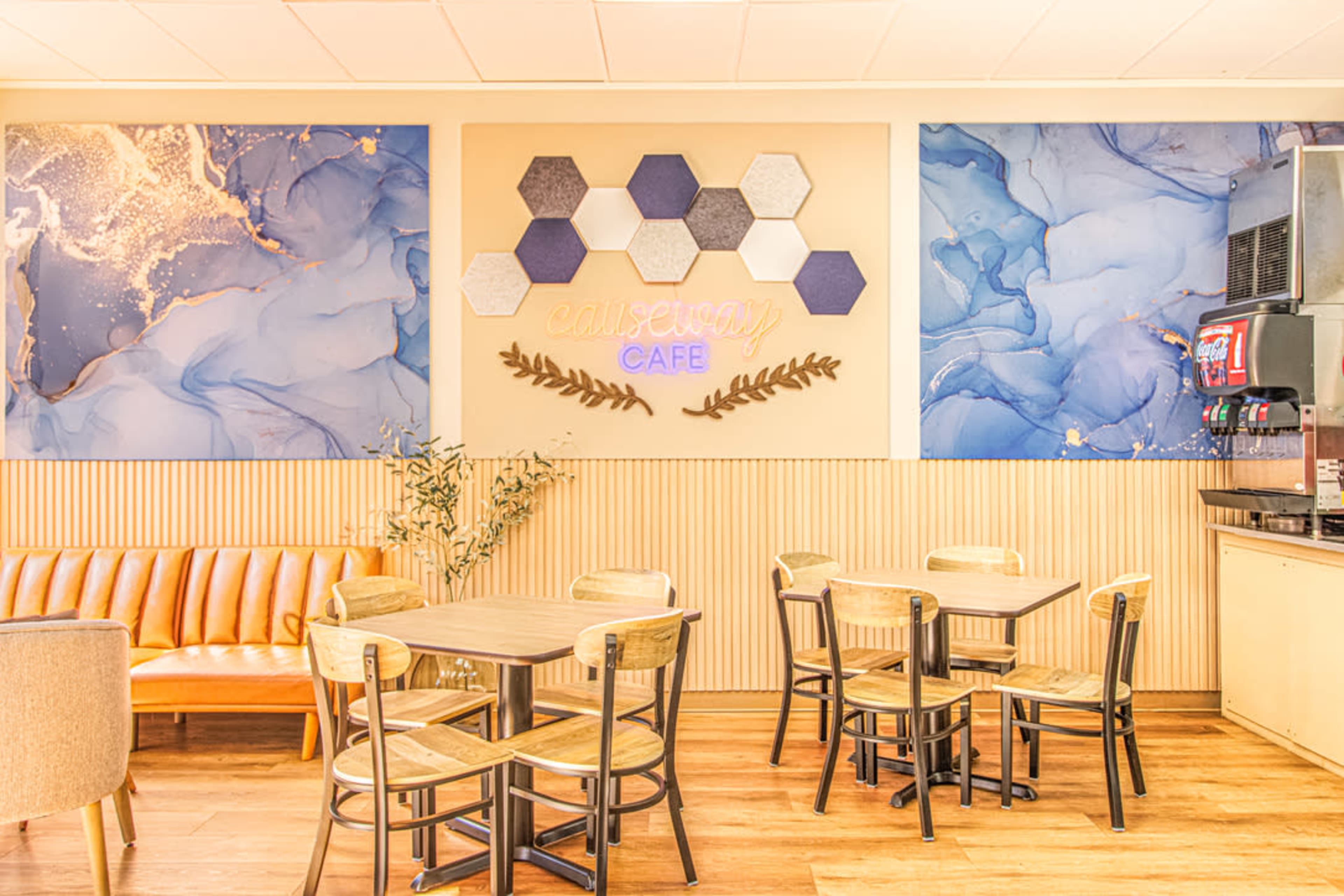 The image shows a café interior featuring a brown leather couch, wooden tables and chairs, and a decorative wall with hexagonal shapes and blue abstract murals.