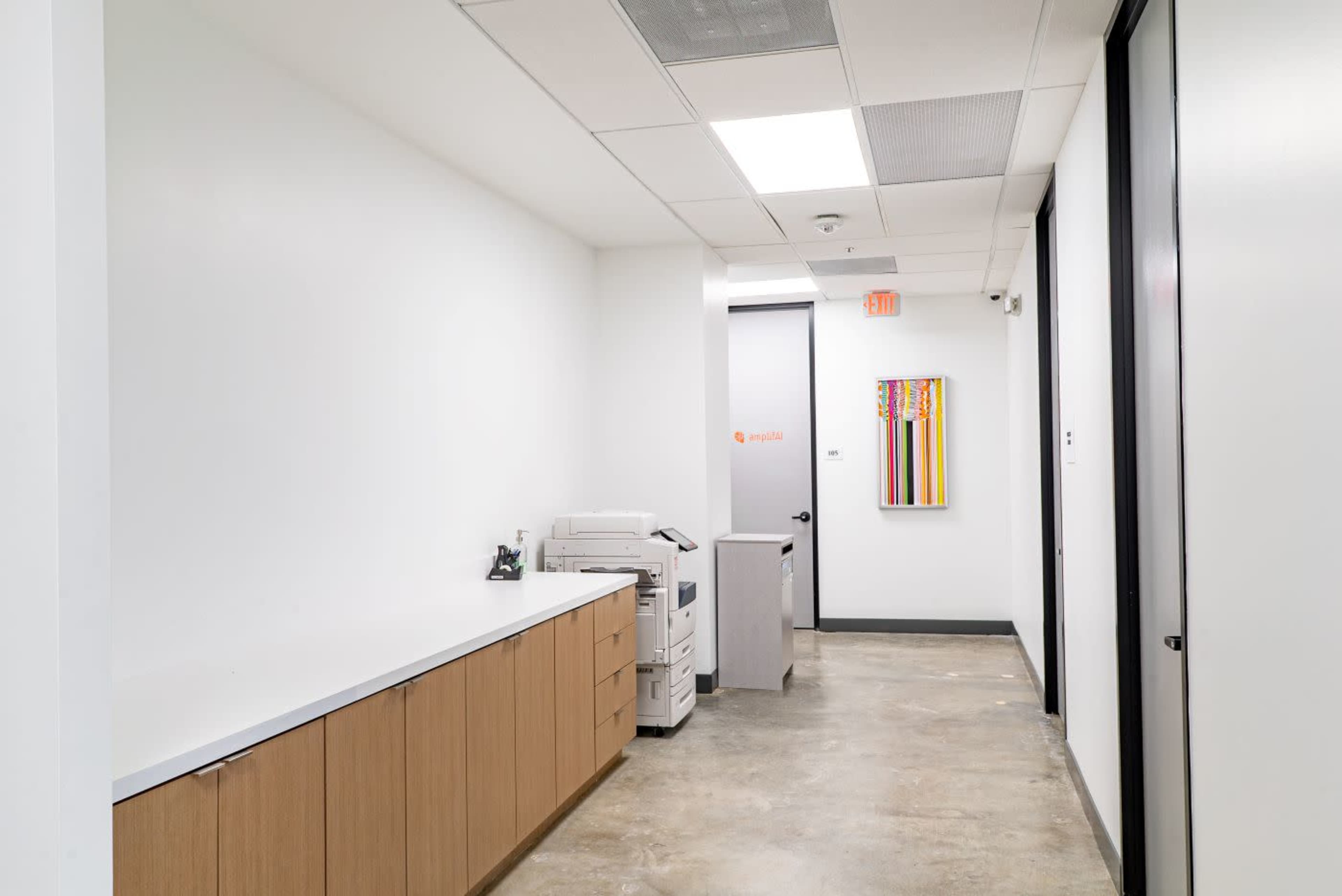 The image shows a narrow hallway in an office building, featuring a beige counter with office machines and a door at the end.