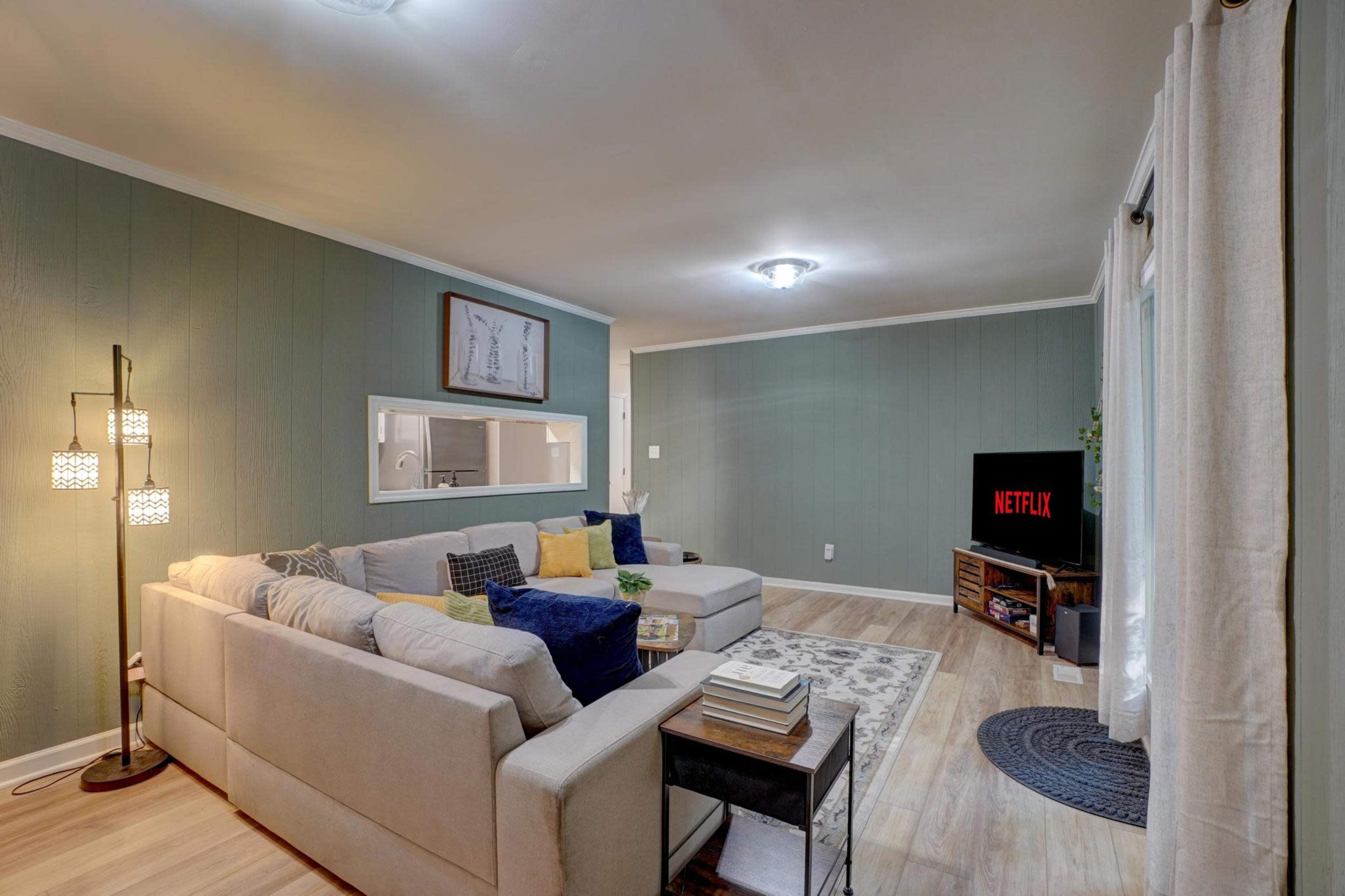 A modern living room features a light-colored sectional sofa, a television displaying the Netflix logo, and wall decorations against a green-paneled wall.