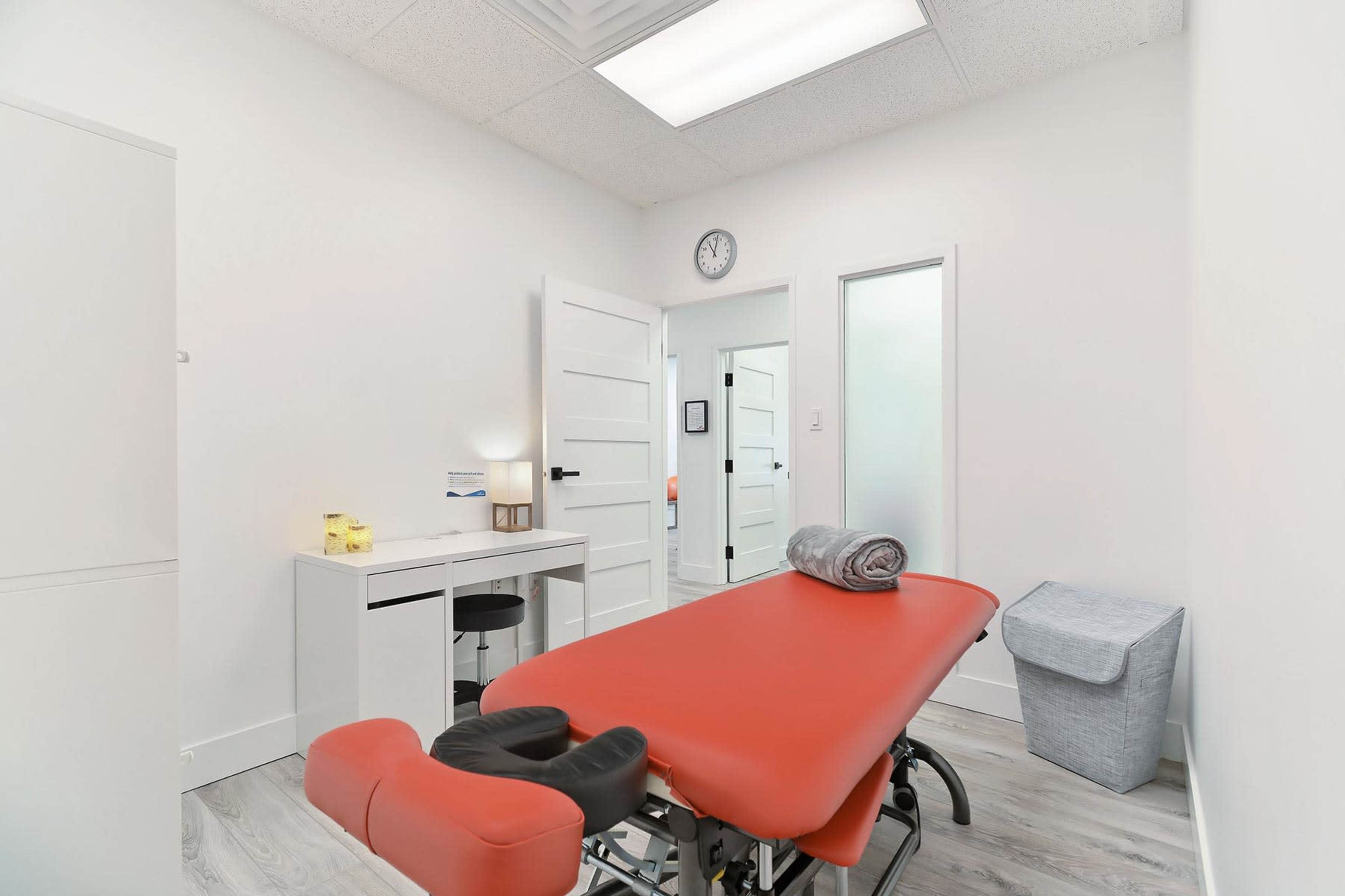 A treatment room with an orange examination table, a desk, and a waste bin.