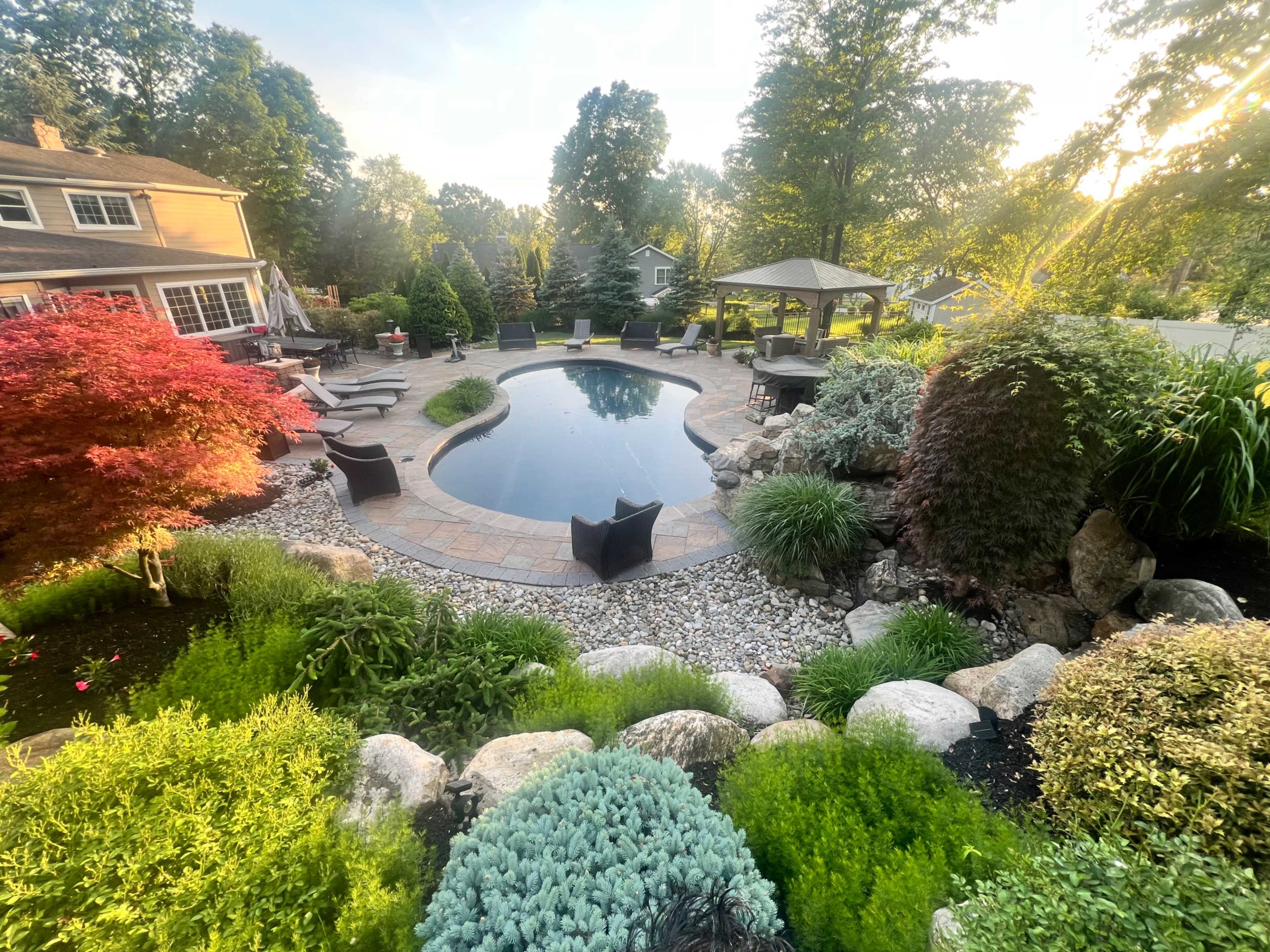A well-landscaped backyard features a swimming pool surrounded by stone pathways, lush greenery, and patio seating areas.