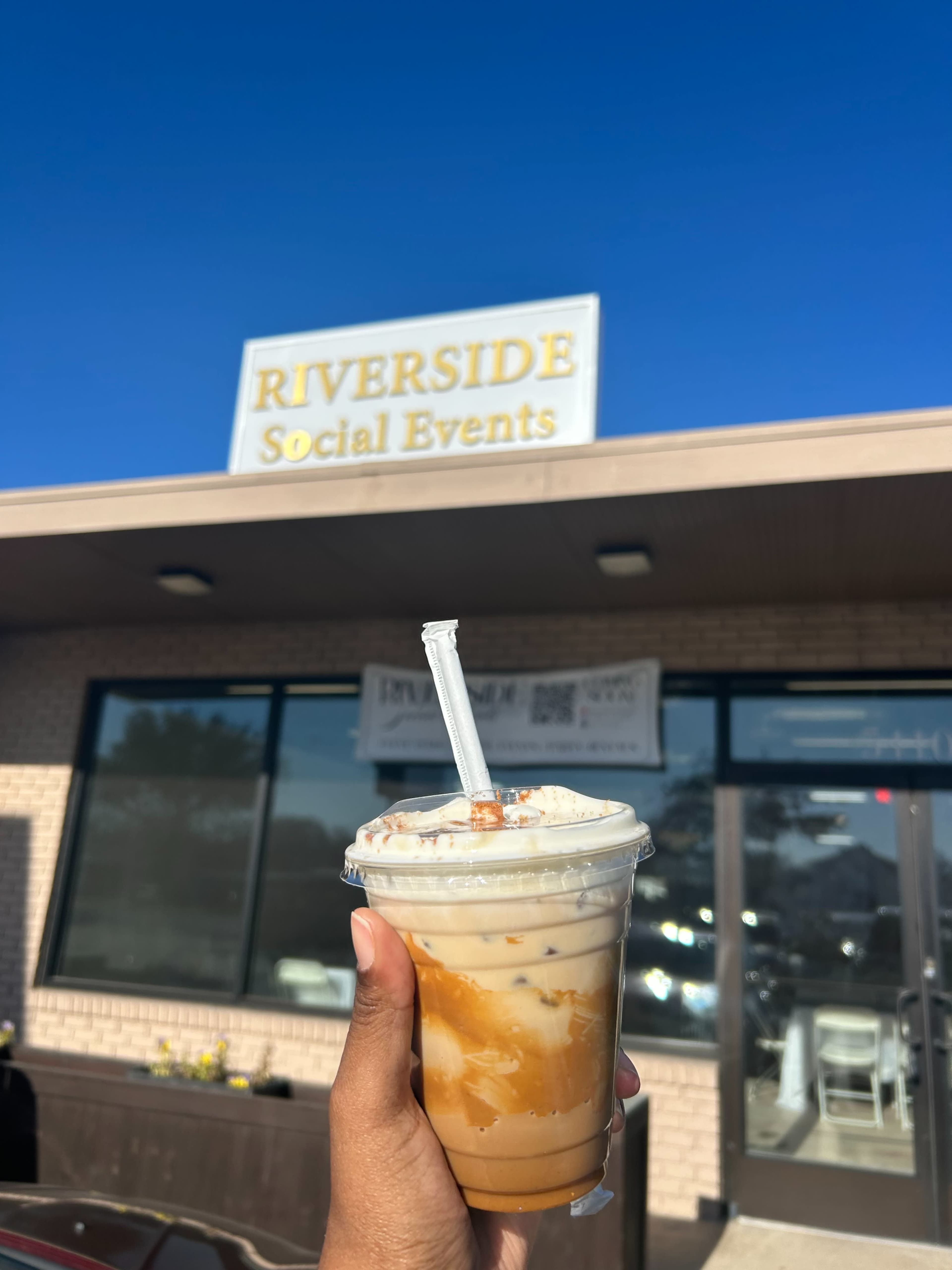 A person holds a cup of iced coffee in front of a building with a sign that reads "RIVERSIDE Social Events" under a clear blue sky.