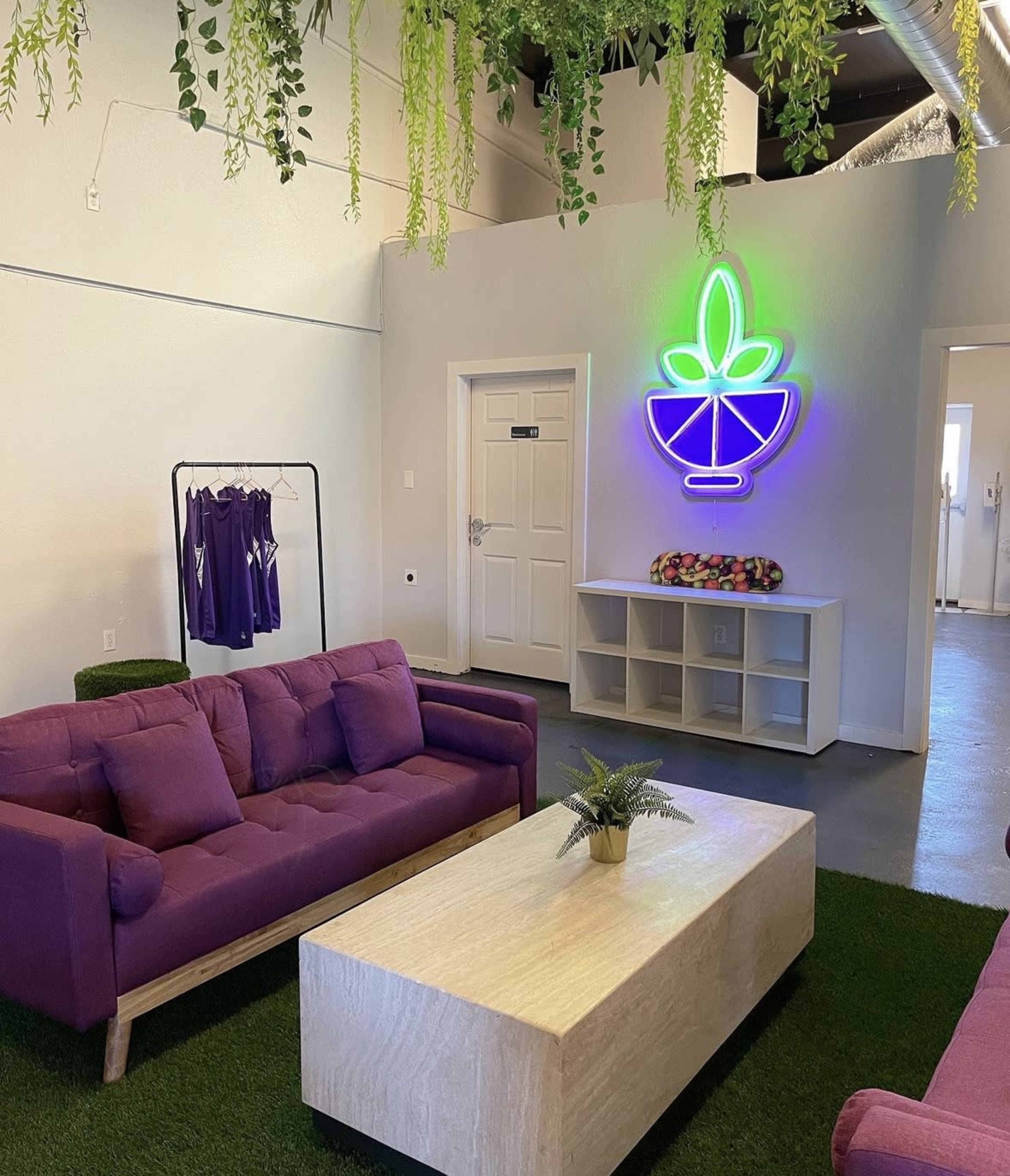 A bright interior space features purple sofas, a wooden coffee table, and a neon sign of a plant, surrounded by greenery and clothing hanging on a rack.