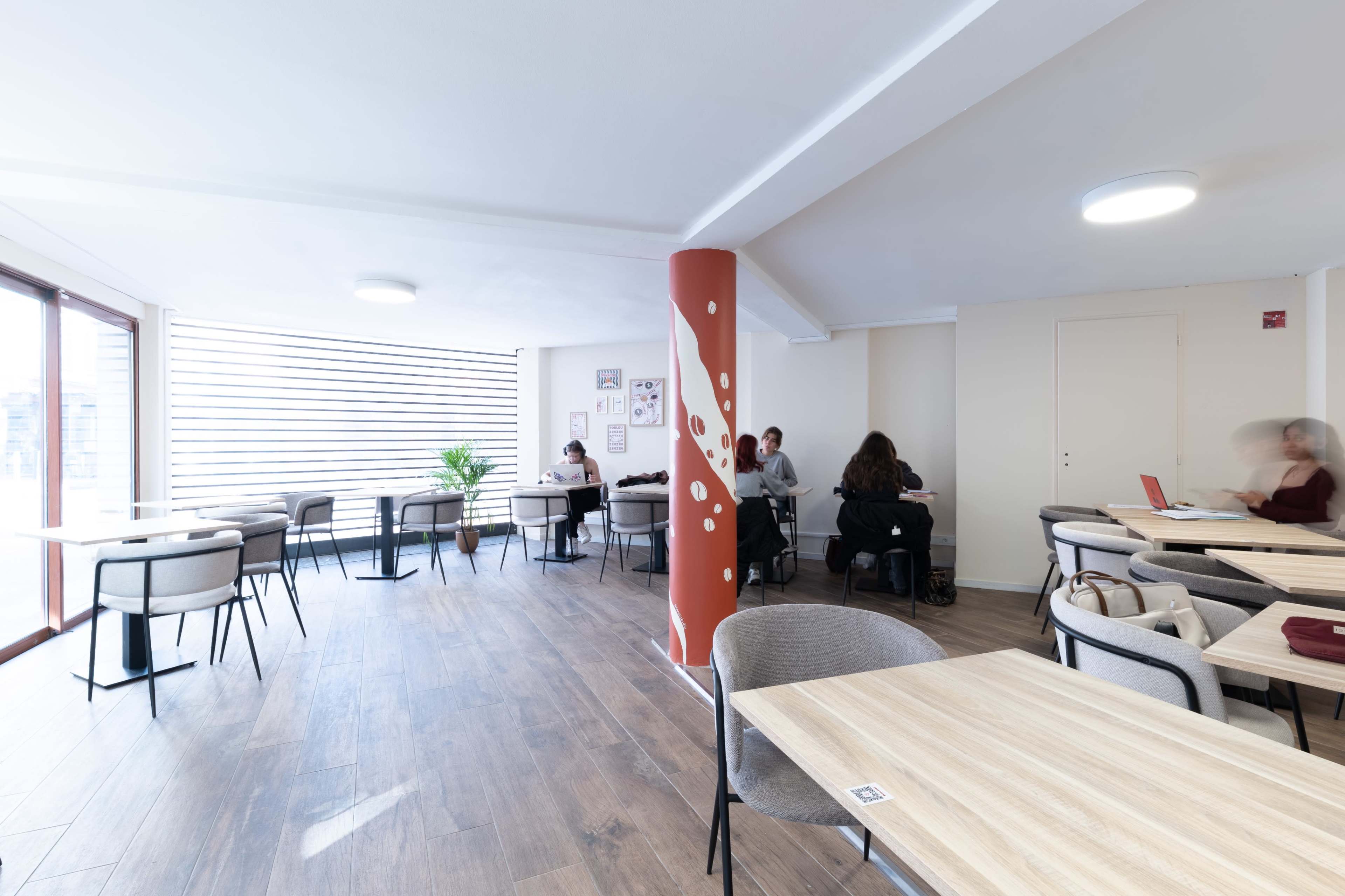 The image shows a spacious cafe with several tables and chairs, featuring a central column with a decorative design, while a few patrons are seated and engaged in activities.