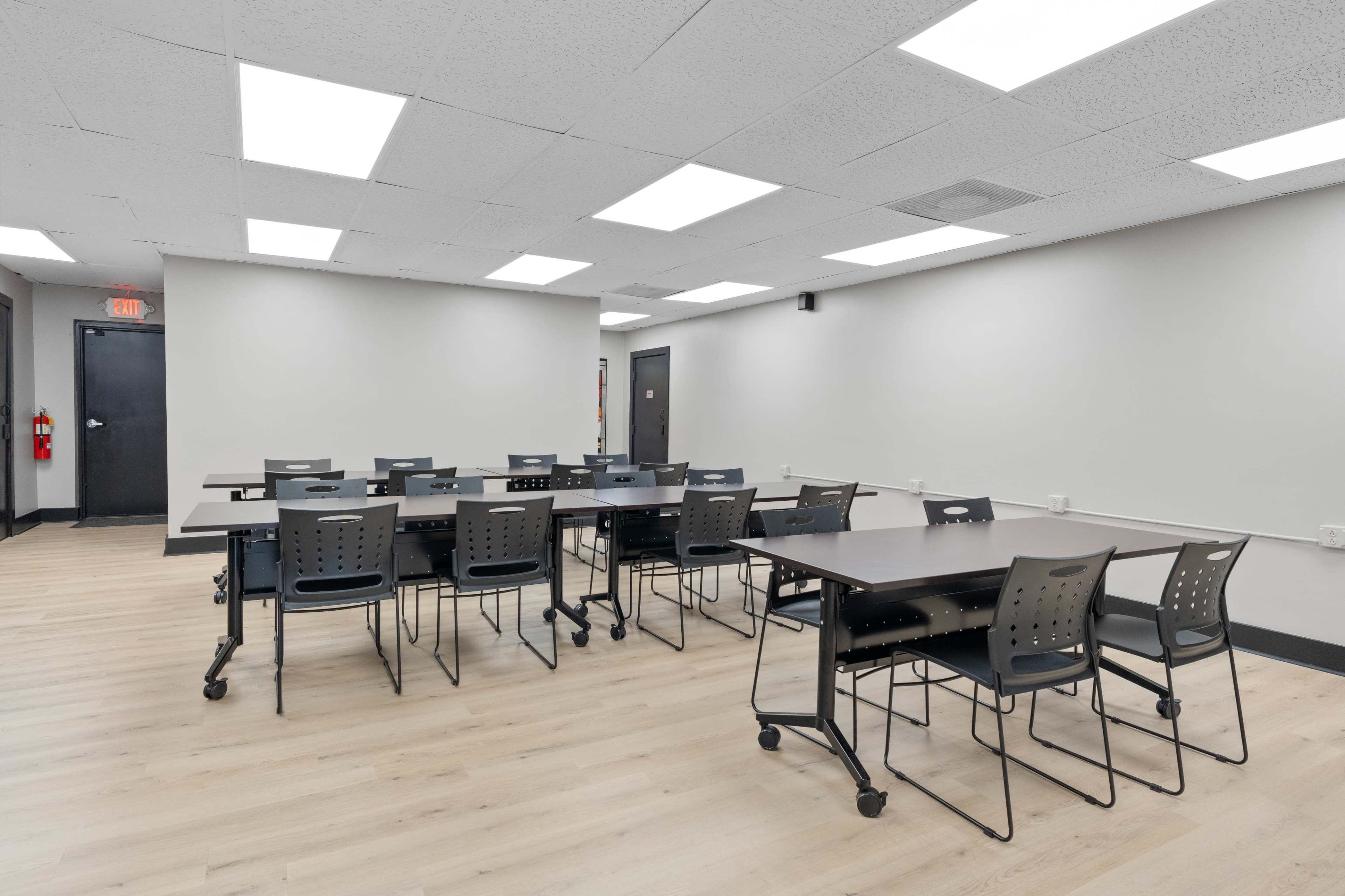 A classroom features multiple black tables and chairs arranged neatly on a light-colored wooden floor, with bright overhead lights illuminating the space.
