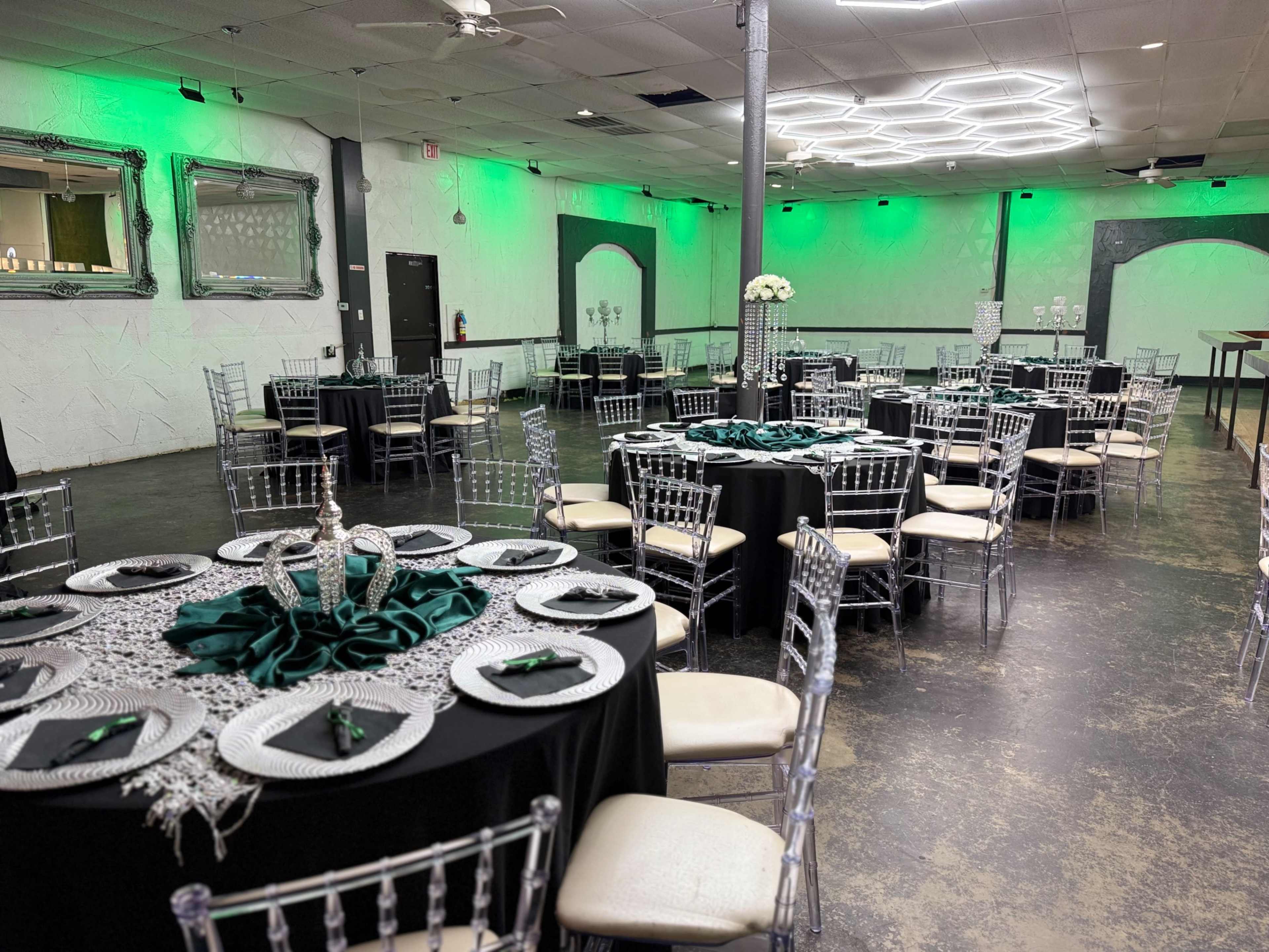 The image depicts a banquet hall set up for an event, featuring round tables covered with black linens and adorned with green accents, surrounded by transparent chairs.