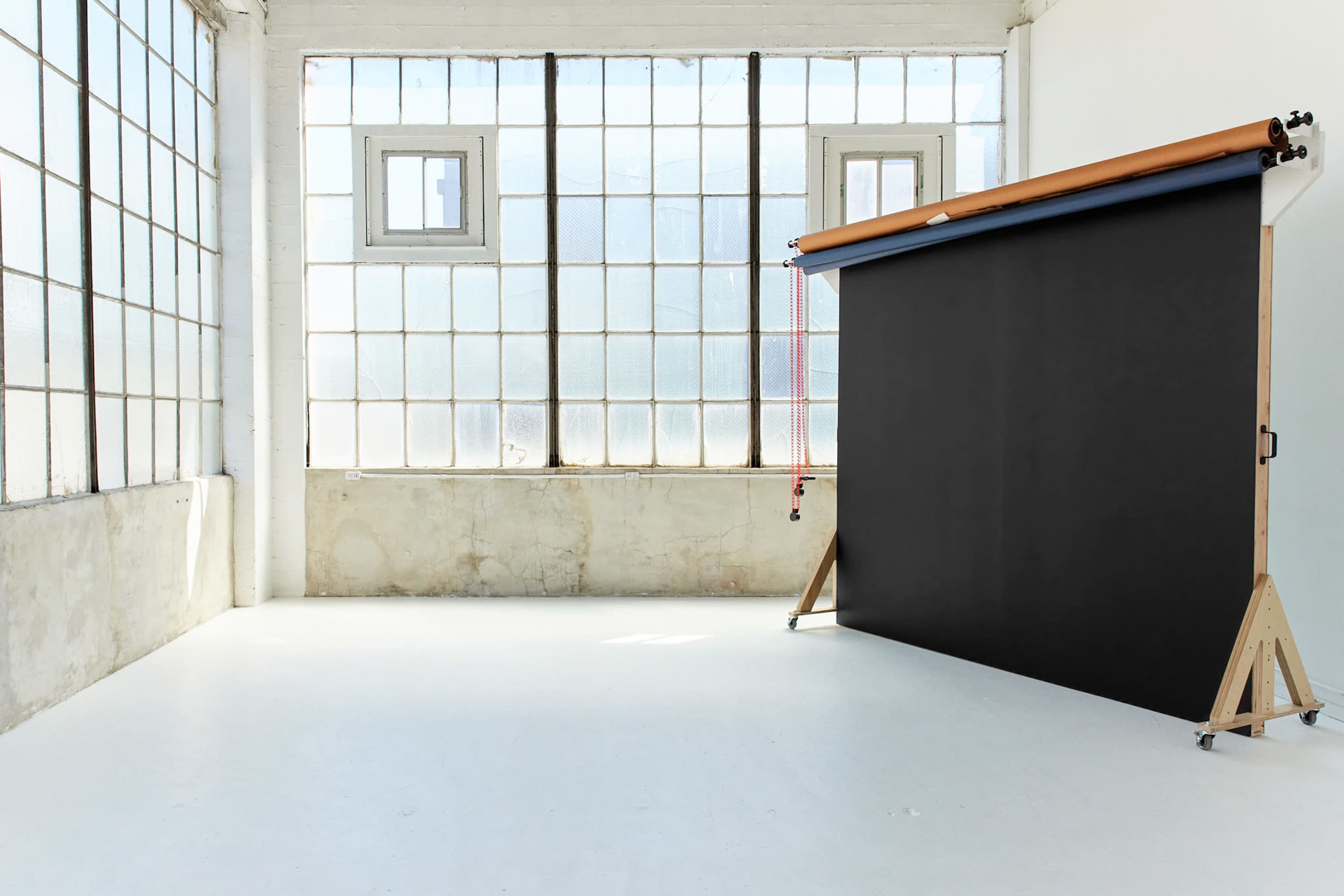 The image shows a spacious, well-lit photography studio featuring large windows and a black backdrop on a wooden stand.