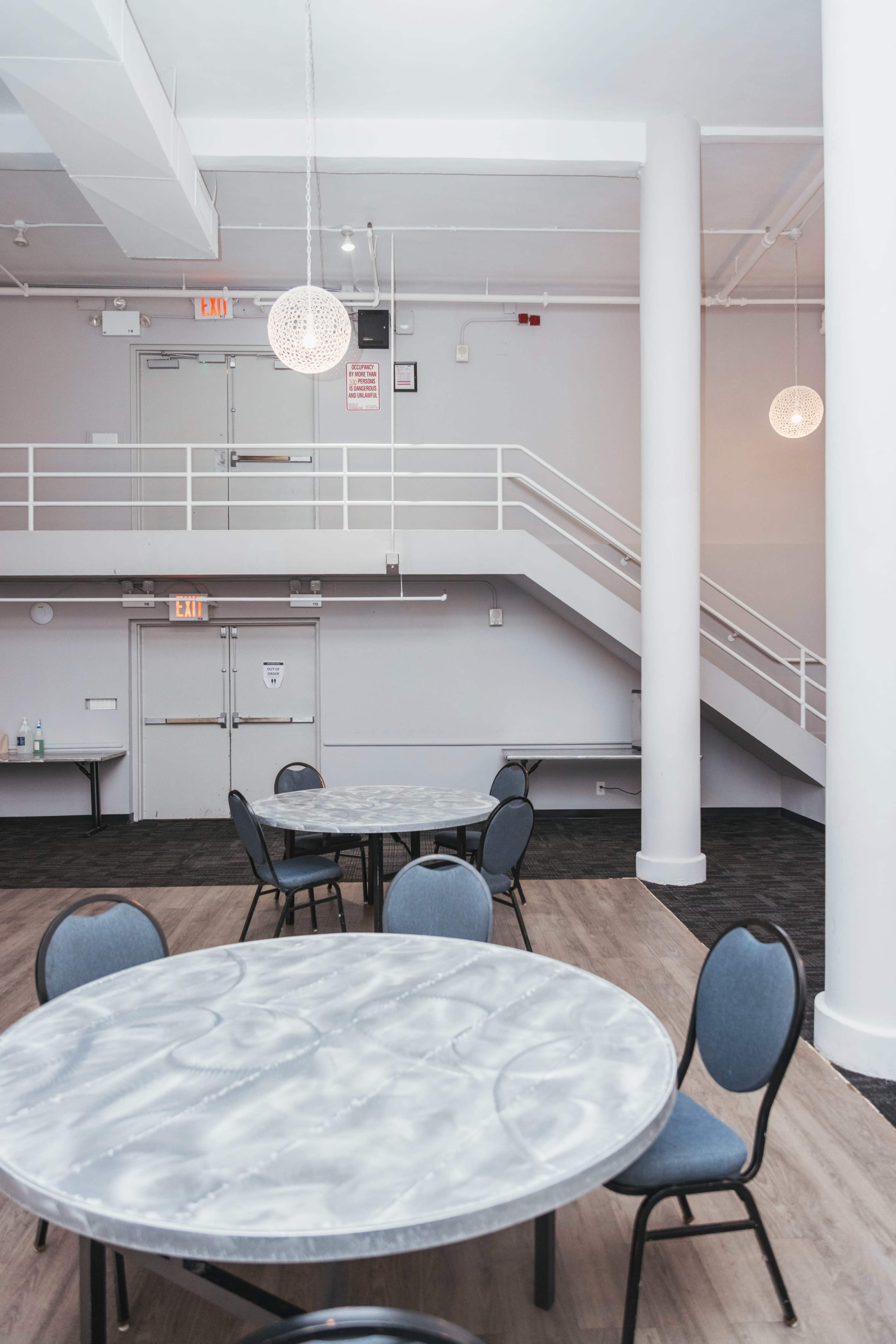 The image shows a modern, spacious event room with several round tables and a staircase in the background.
