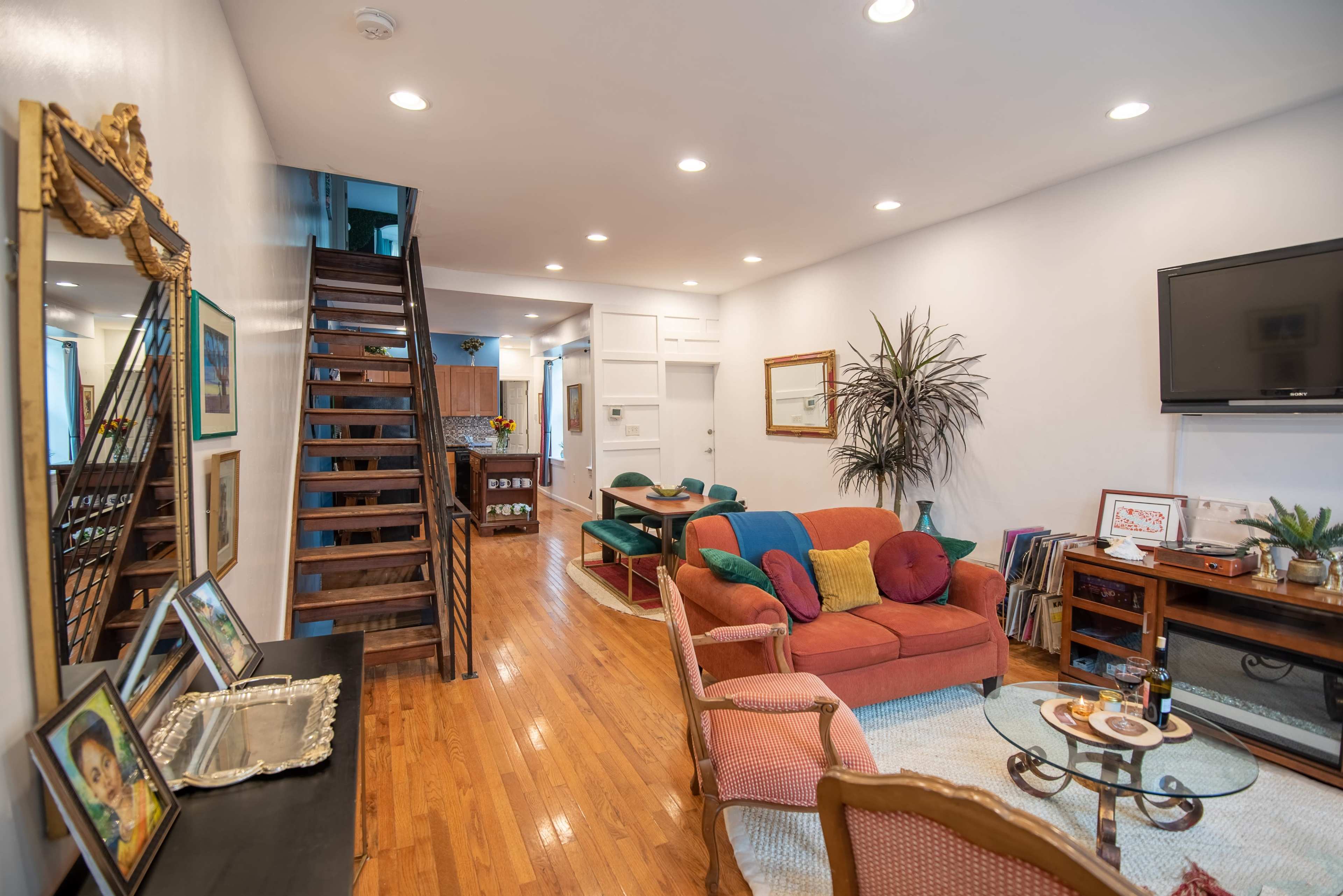 The image shows a living room with a staircase, orange couches, a wooden floor, and a television mounted on the wall.