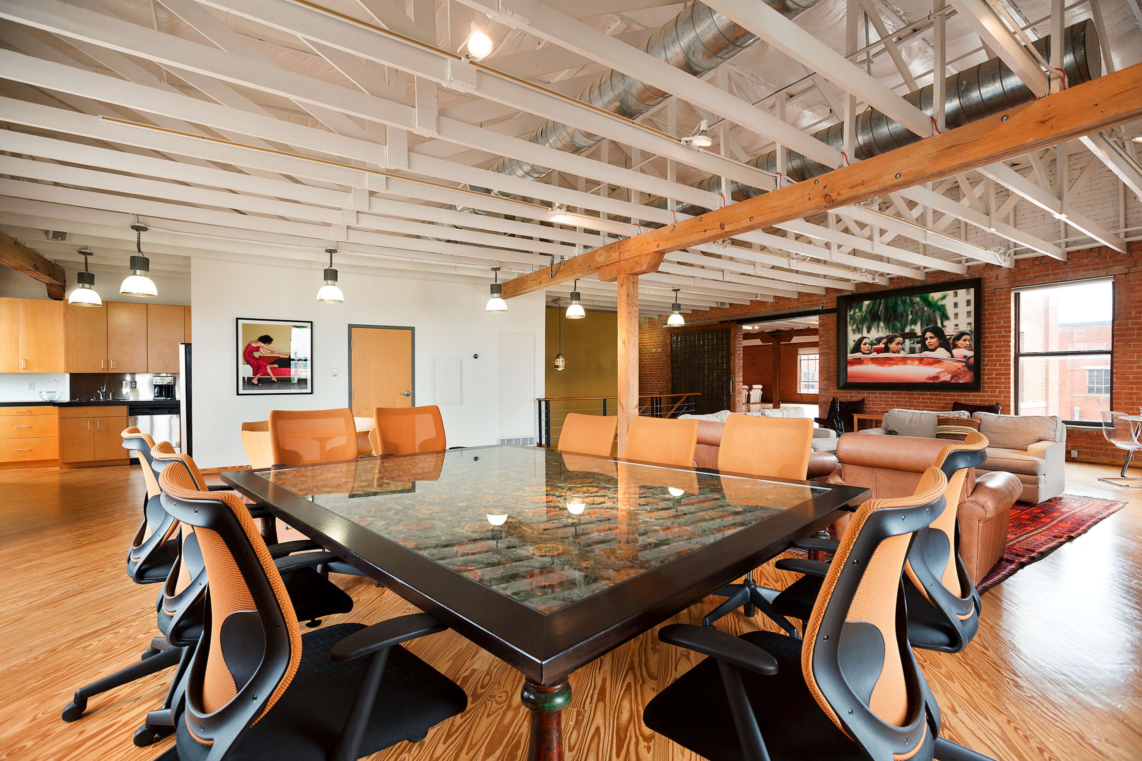 A modern office space with a large glass conference table surrounded by ergonomic chairs, hardwood flooring, and exposed beams, featuring a lounge area with couches and artwork on the walls.