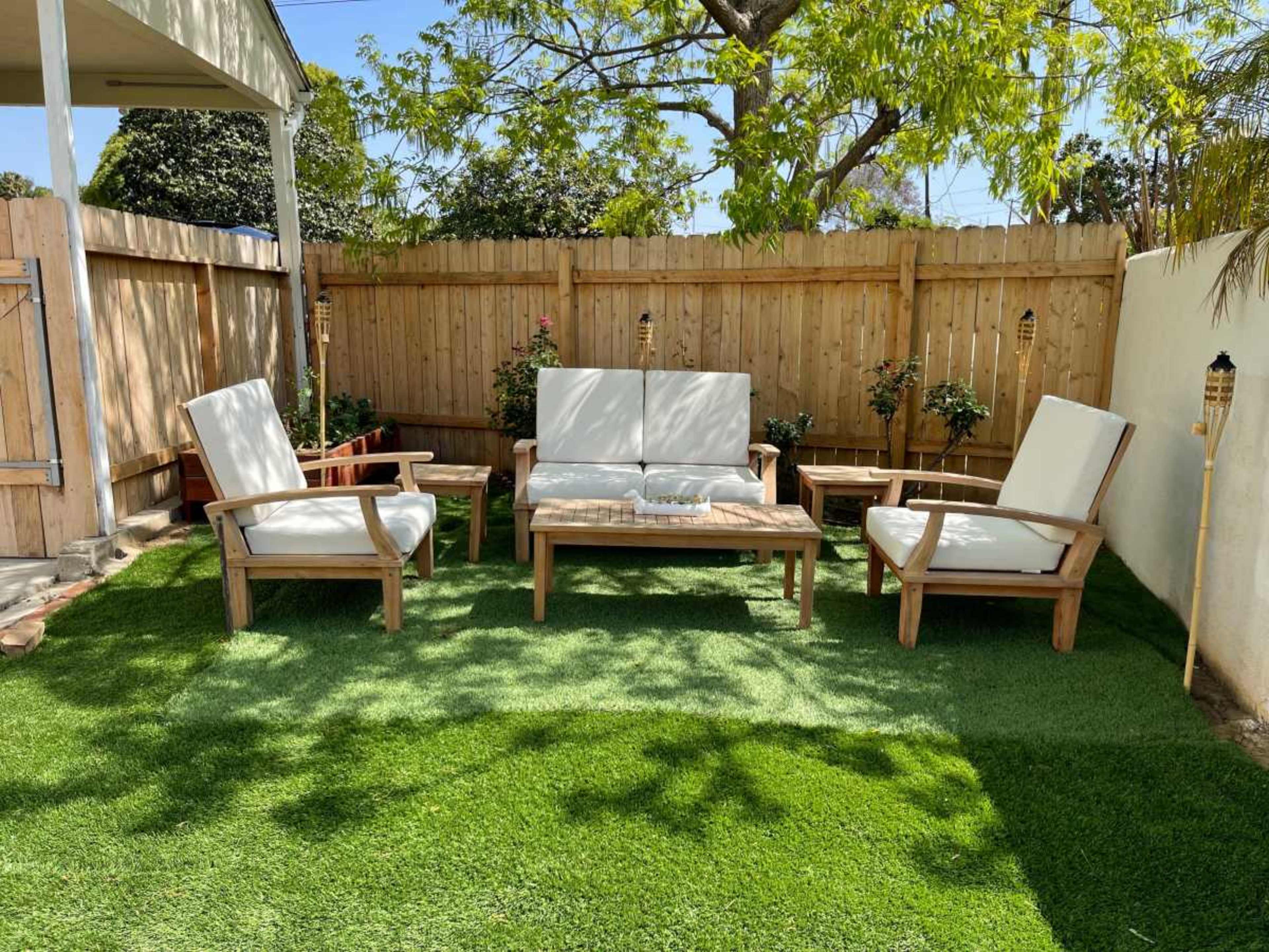 A seating area with wooden furniture and white cushions is situated on artificial grass within a fenced backyard.