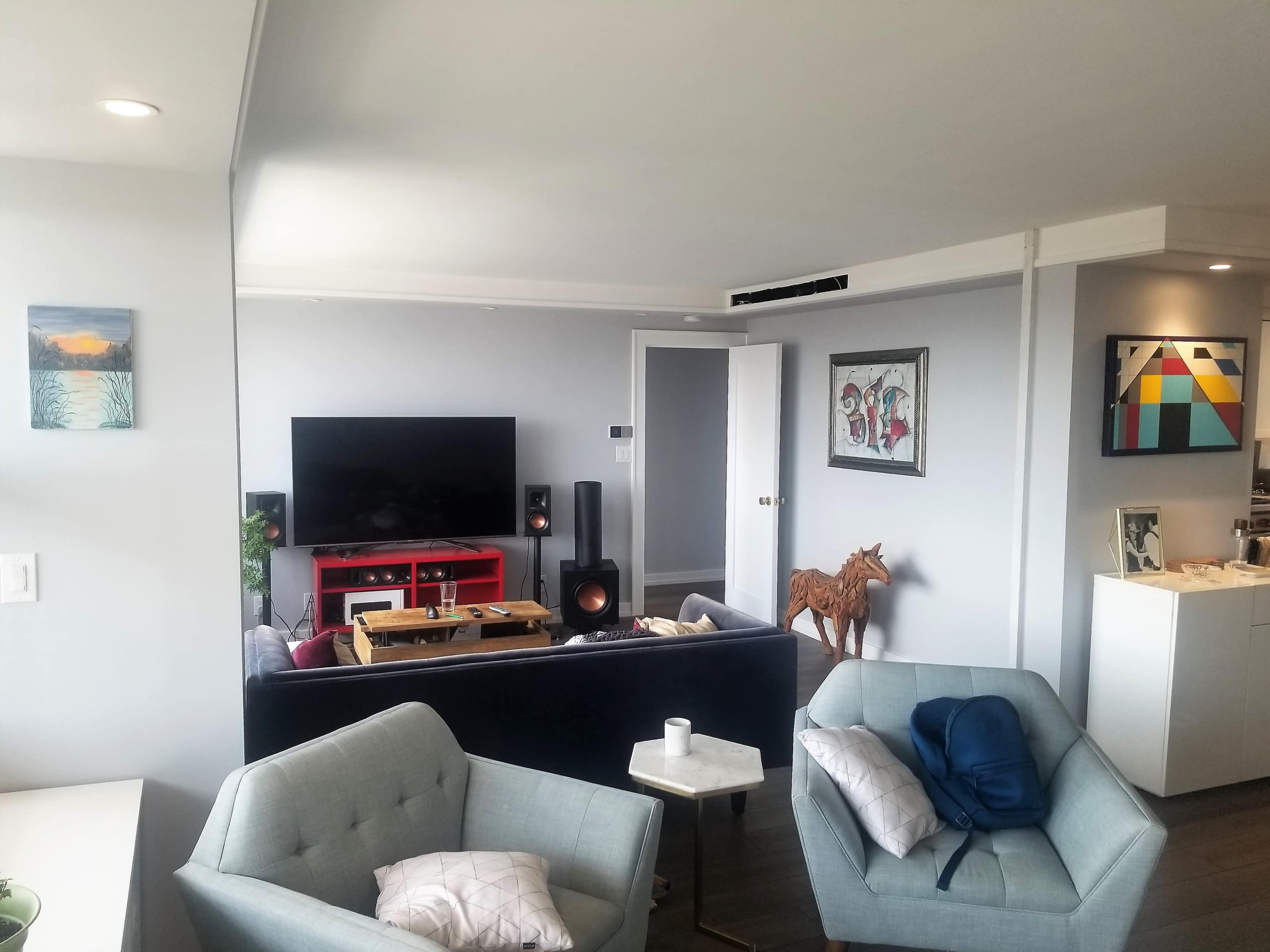 A modern living room with a television set on a red stand, a dark sofa, and two light blue armchairs, all accented by various artworks on the walls.