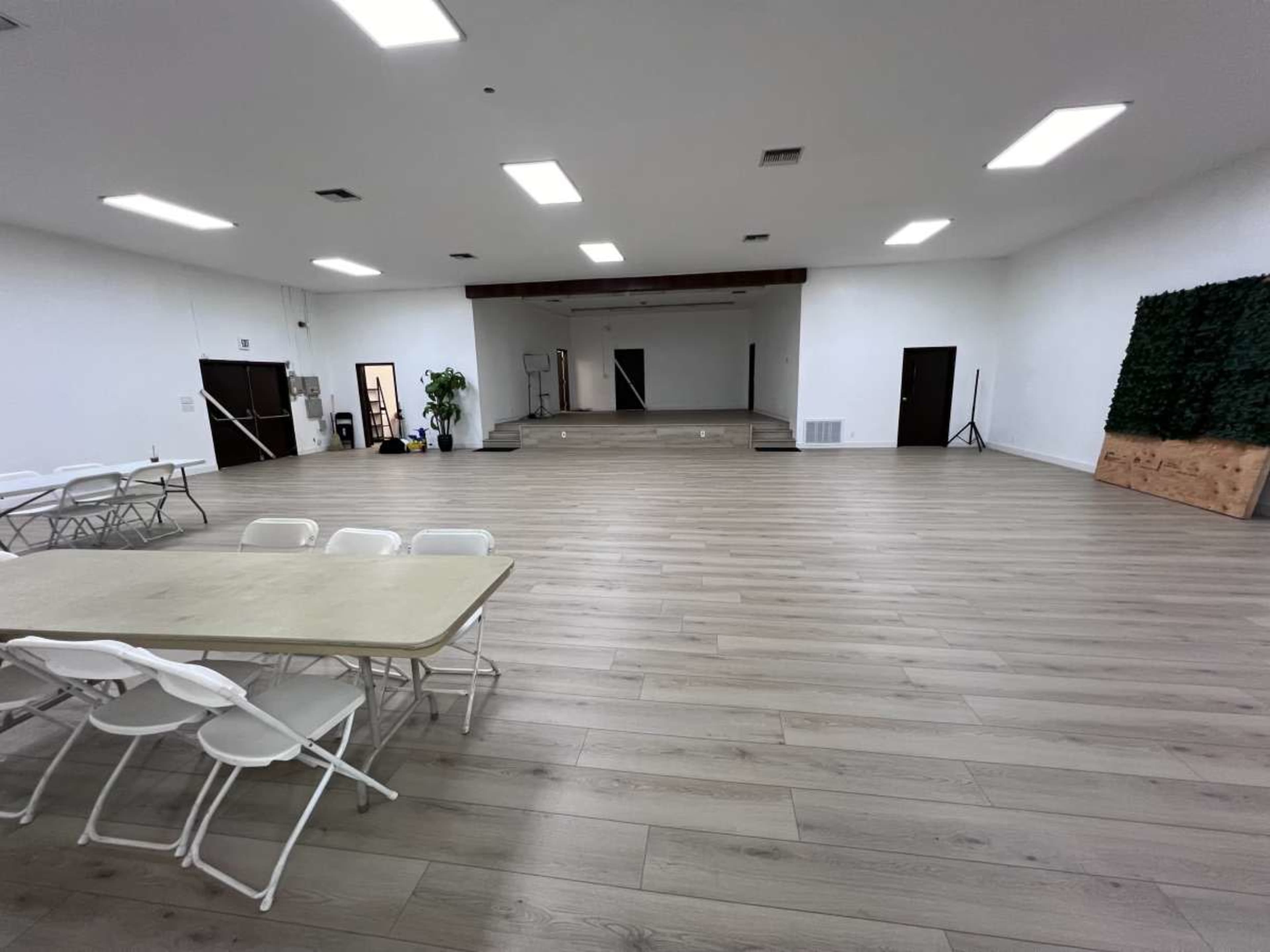 The image shows a large, empty room with wooden flooring, featuring a stage at one end and several folding tables set up in the foreground.