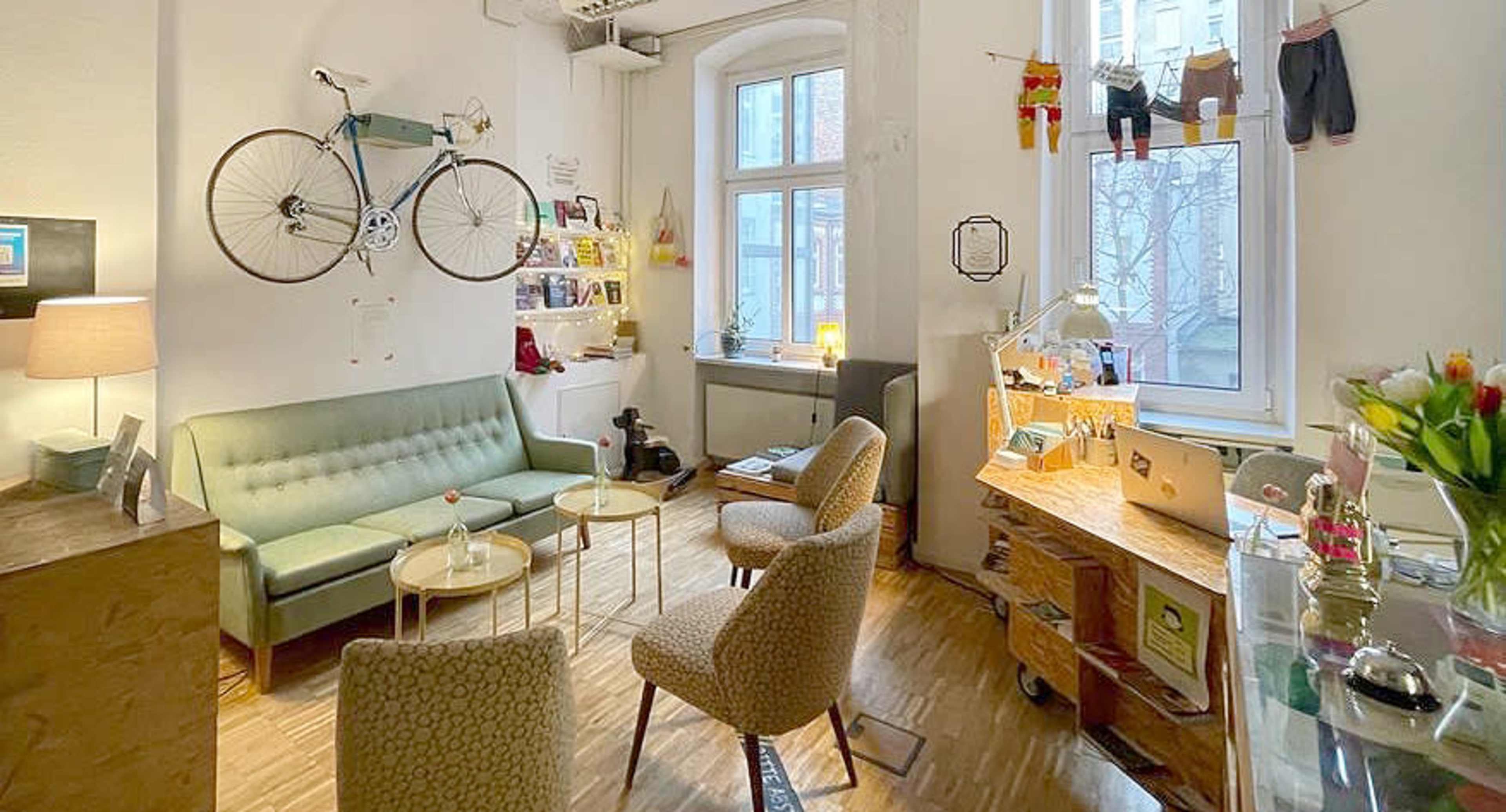 A cozy room with a green sofa, two patterned chairs, a wooden table, a mounted bicycle on the wall, and a small workspace by the window.