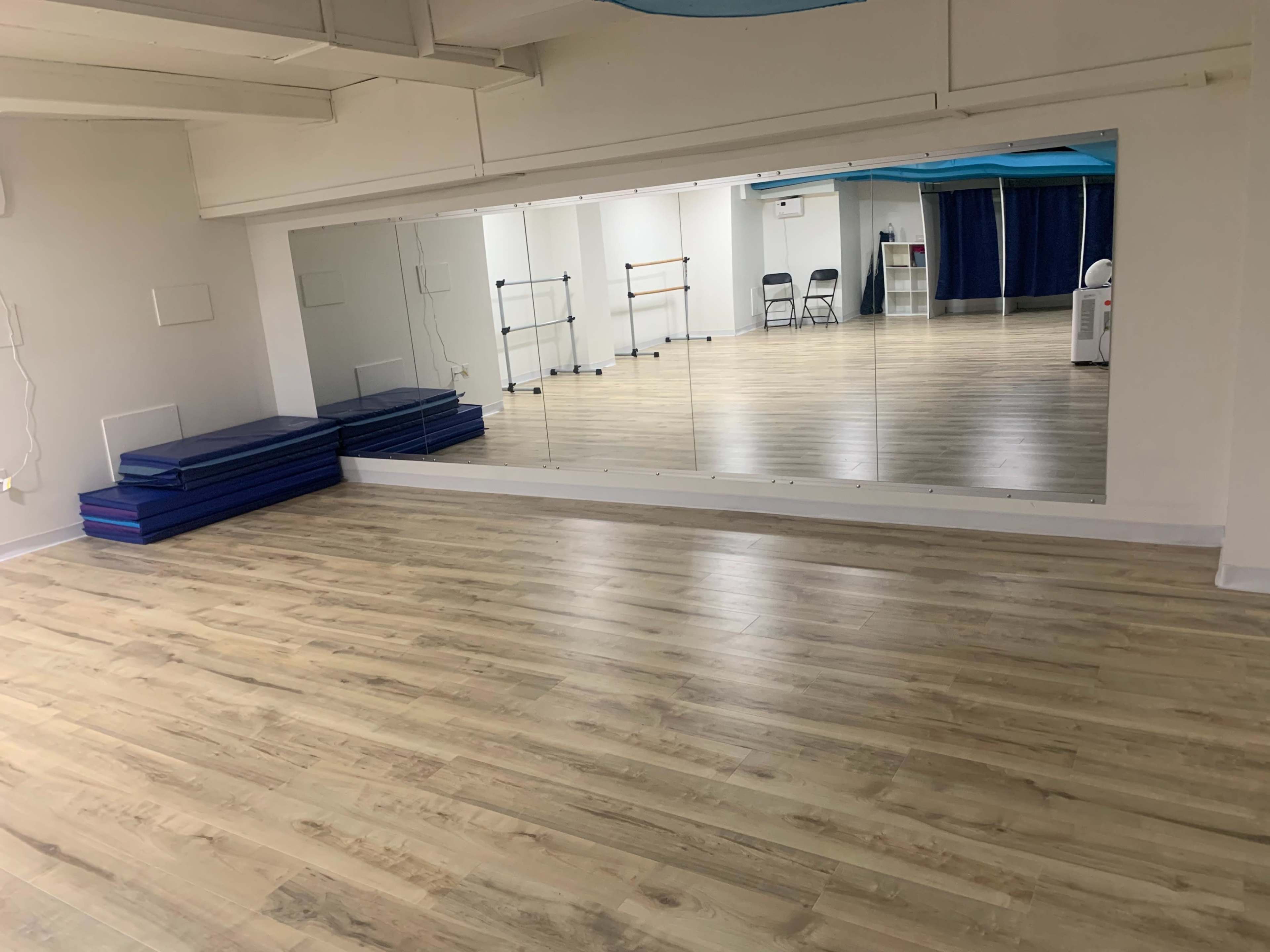 The image shows a bright studio space with wooden flooring, large mirrors on one wall, and exercise mats stacked neatly in the corner.