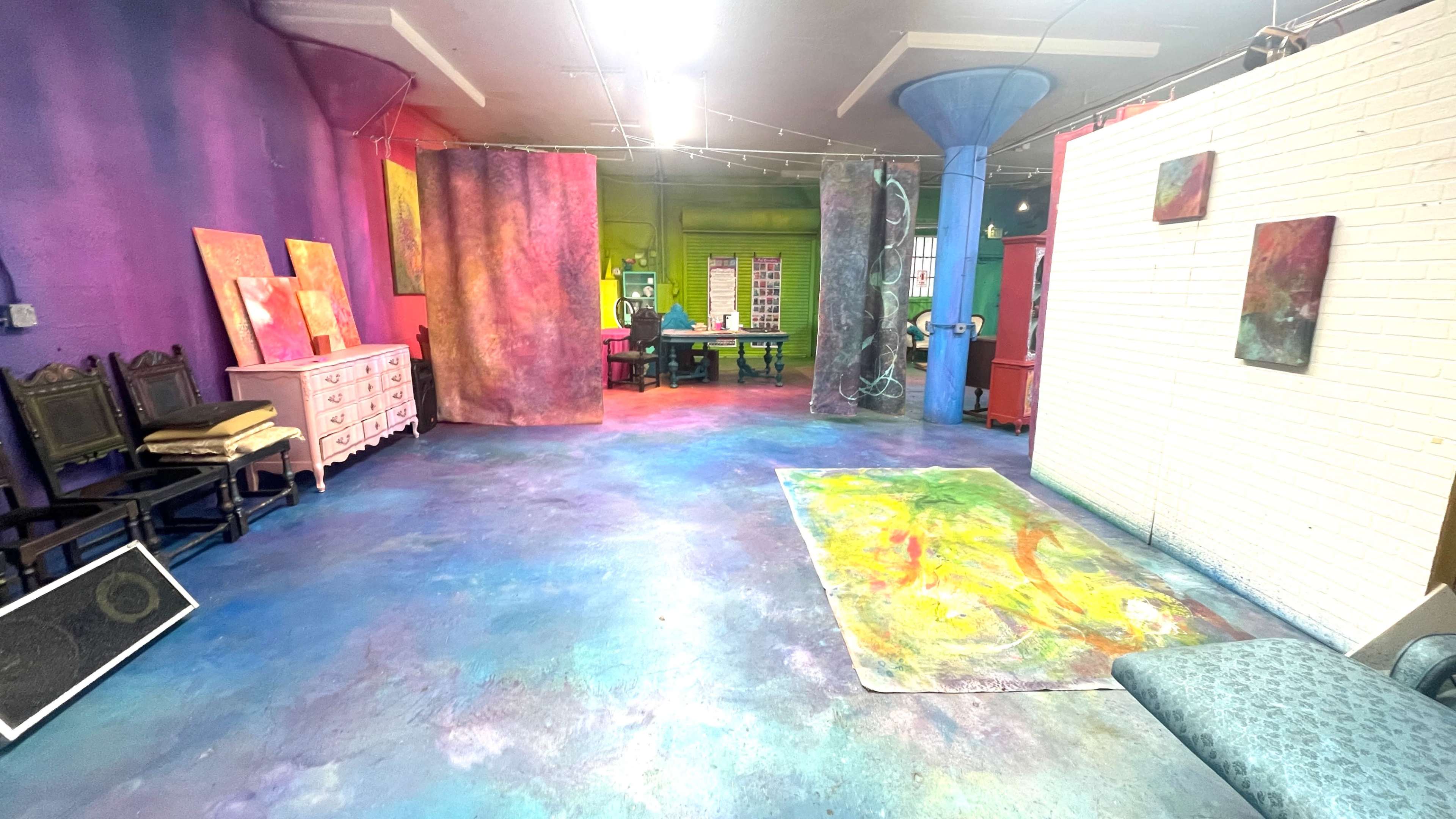The image shows a colorful art studio with vibrant walls, various artworks displayed, and a person working at a table in the background.