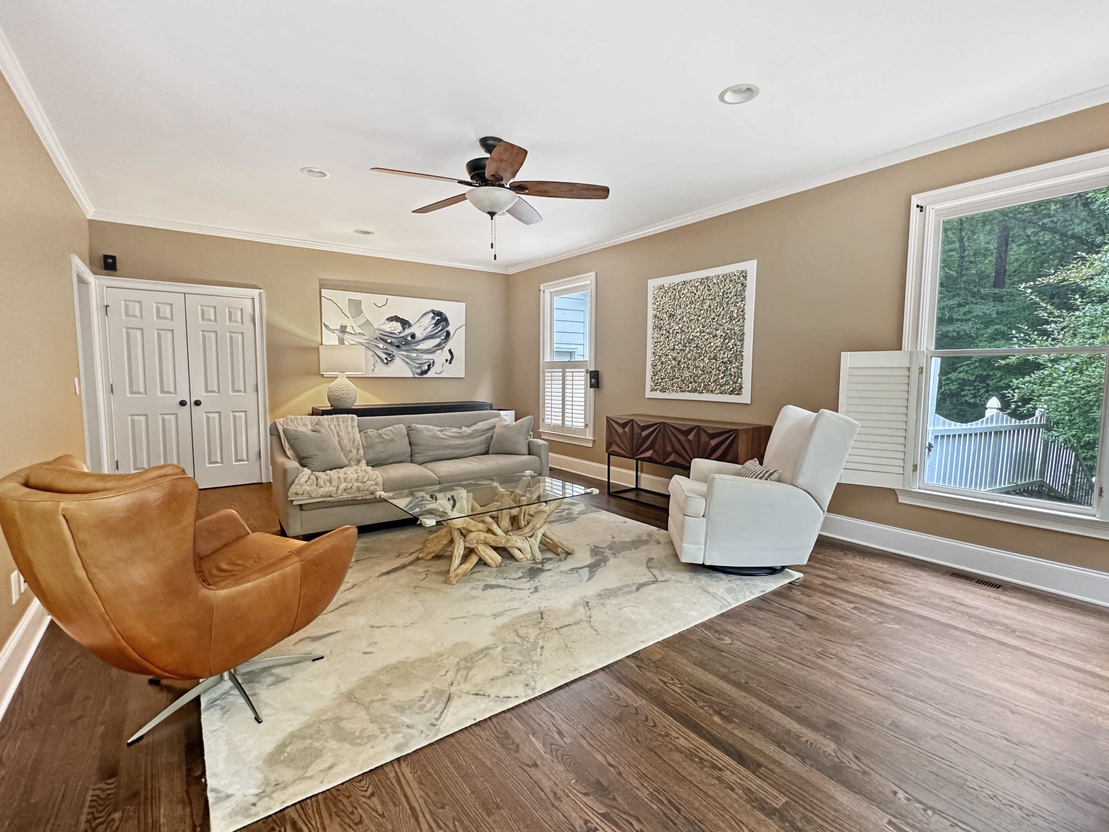 The living room features a beige wall, hardwood floors, a gray sofa, two armchairs, a glass coffee table, and large windows overlooking greenery.
