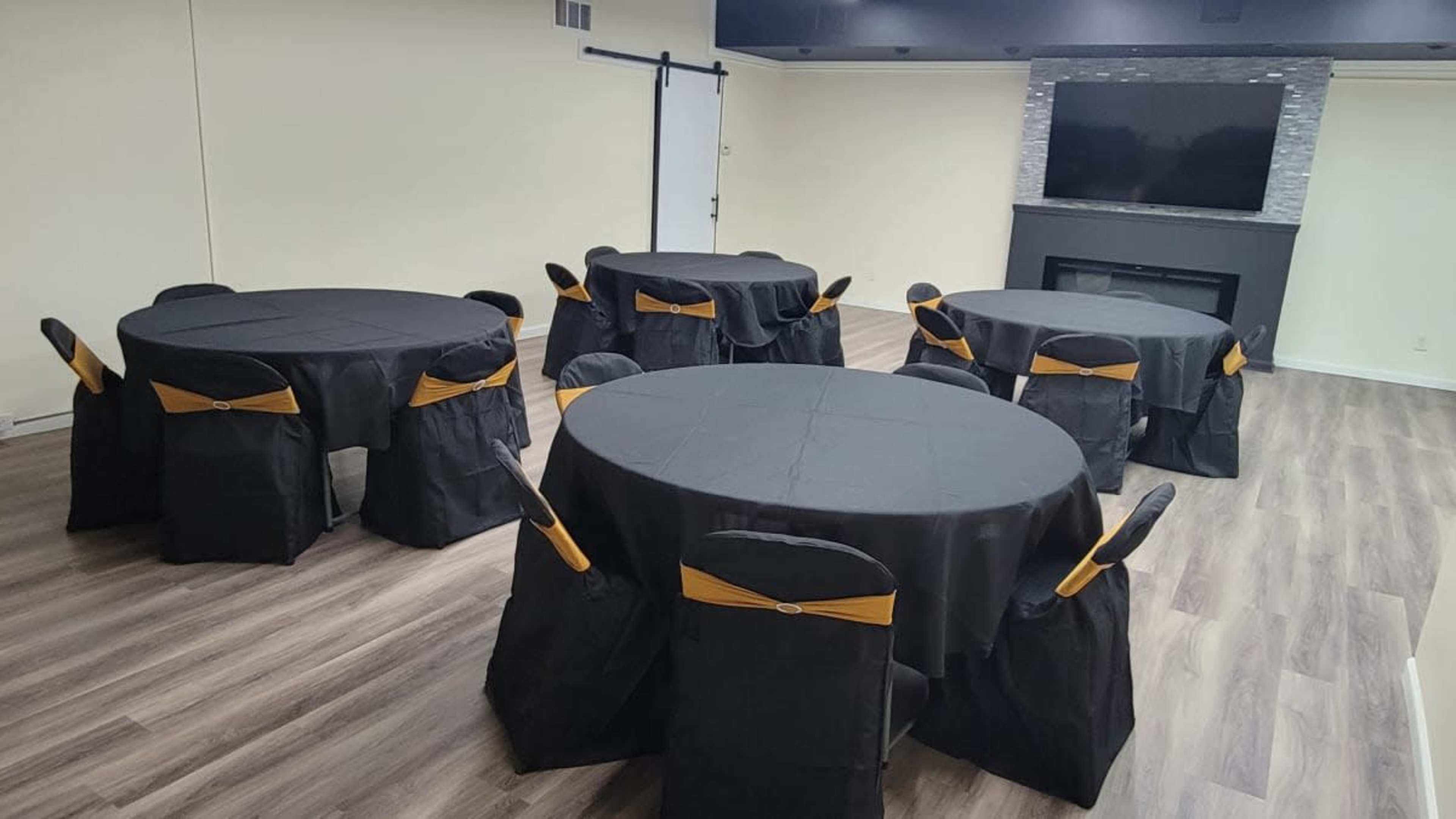 A spacious room features several round tables covered with black tablecloths and adorned with gold sashes on the chairs, along with a stone fireplace and a television on the wall.