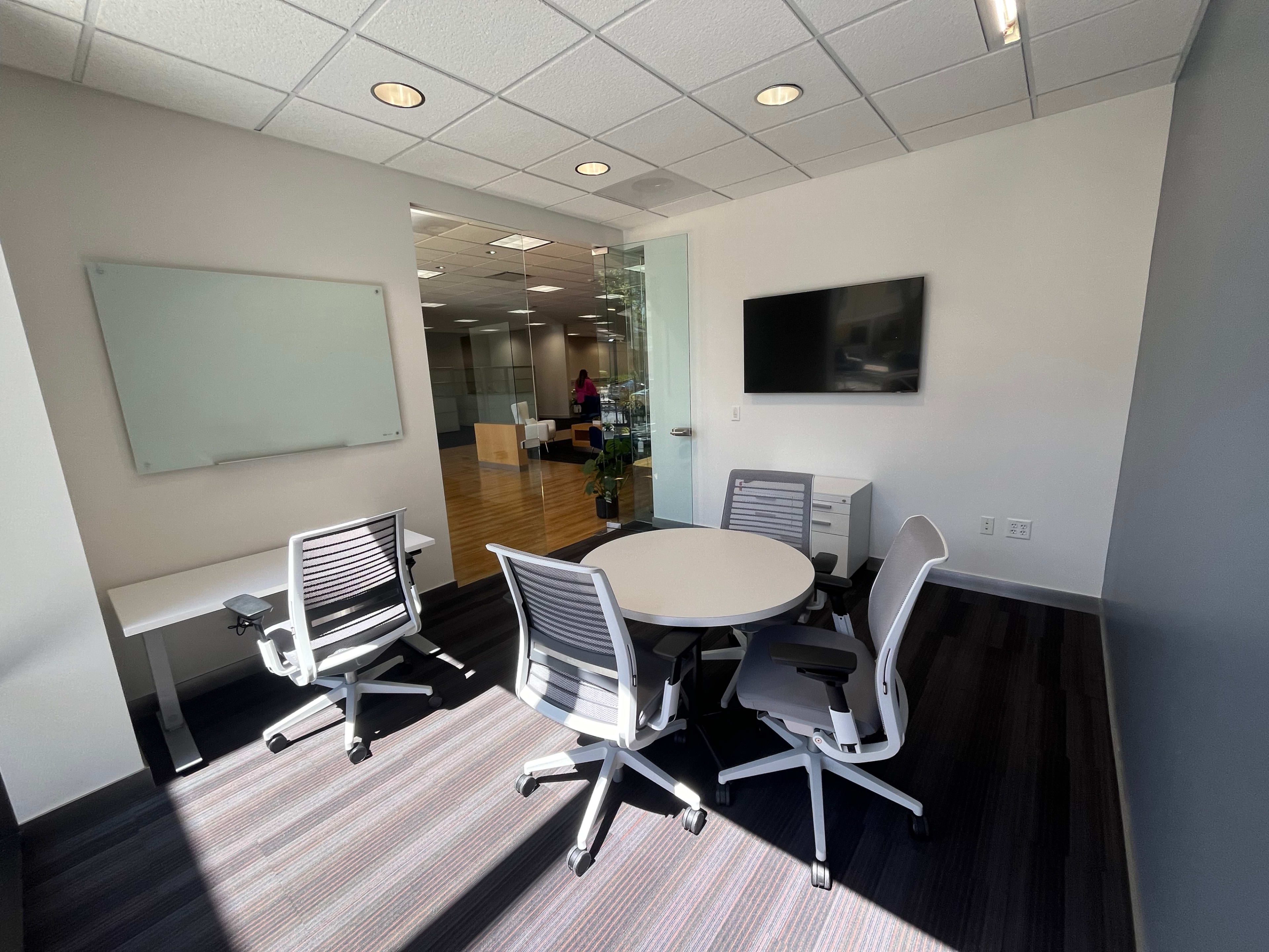 A modern office meeting room with a round table, four office chairs, a whiteboard, and a wall-mounted television.