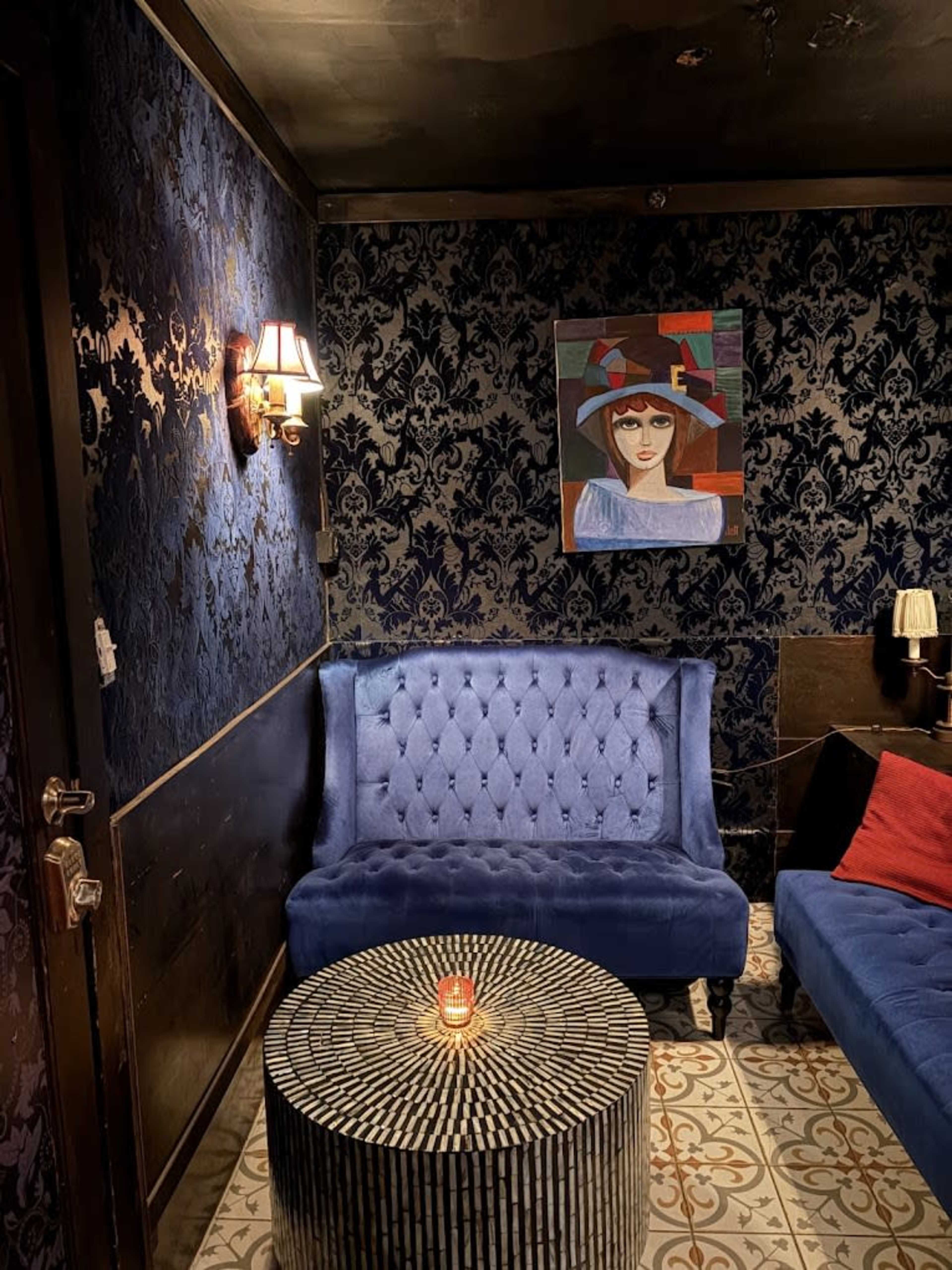 The image shows a cozy lounge area with blue upholstered sofas, a circular striped table, and a wall adorned with a colorful portrait and dark floral wallpaper.