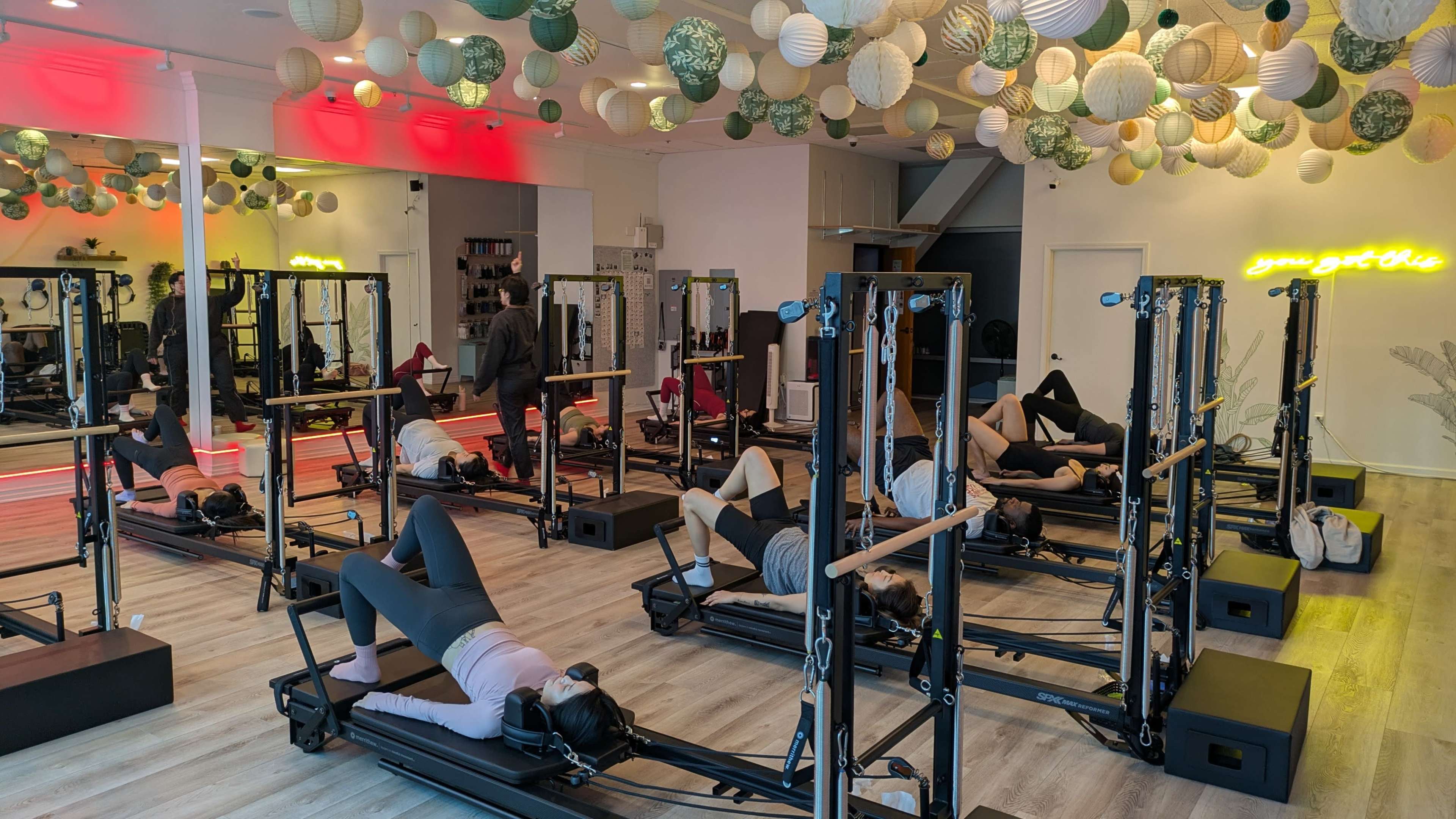 The image shows a fitness studio with several individuals practicing on reformer machines, surrounded by decorative paper lanterns hanging from the ceiling.