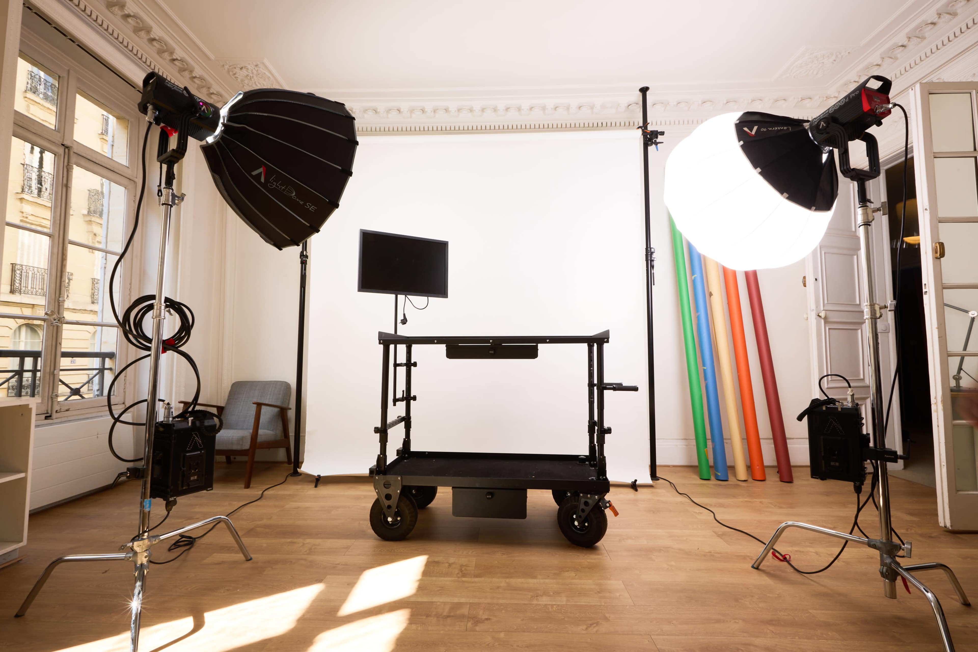 The image shows a photography studio setup with two large light sources, a rolling cart with a monitor, and colorful backdrops leaning against the wall.