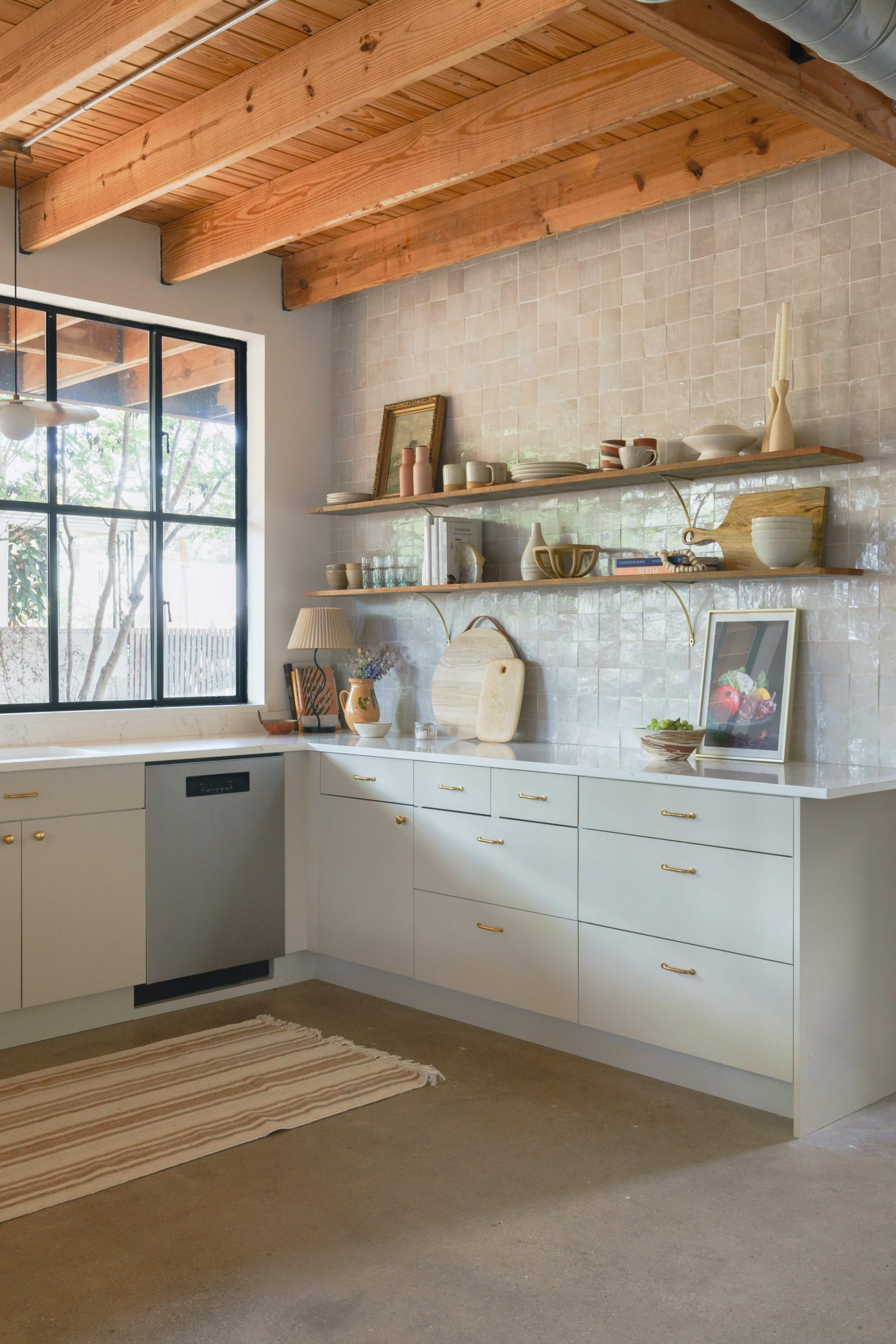 A modern kitchen features a spacious layout with beige cabinetry, open shelving displaying decor, and a large window allowing natural light to enter.