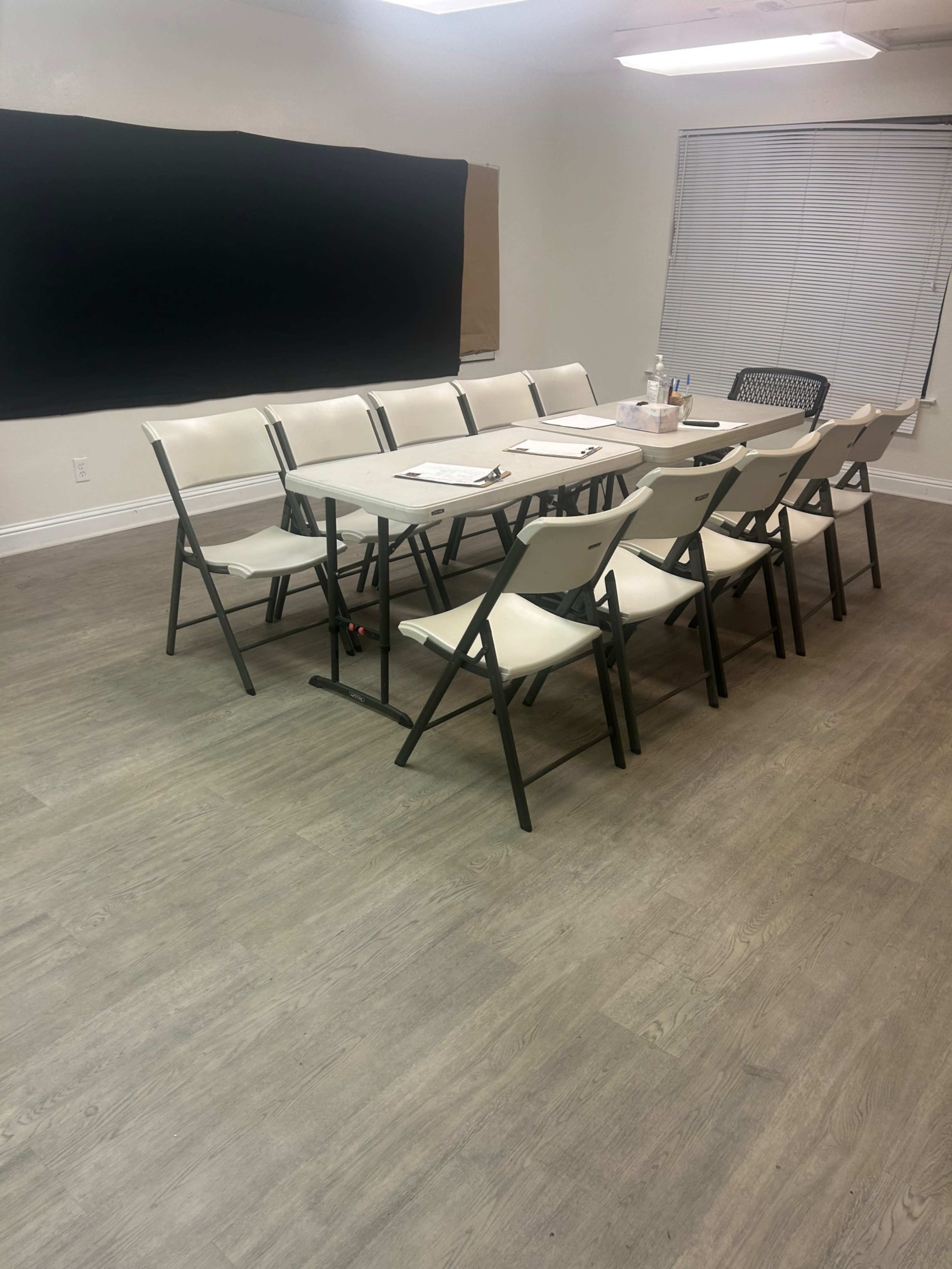 A conference room is set up with a long table surrounded by folding chairs, with a blackboard on one wall and blinds on a window.