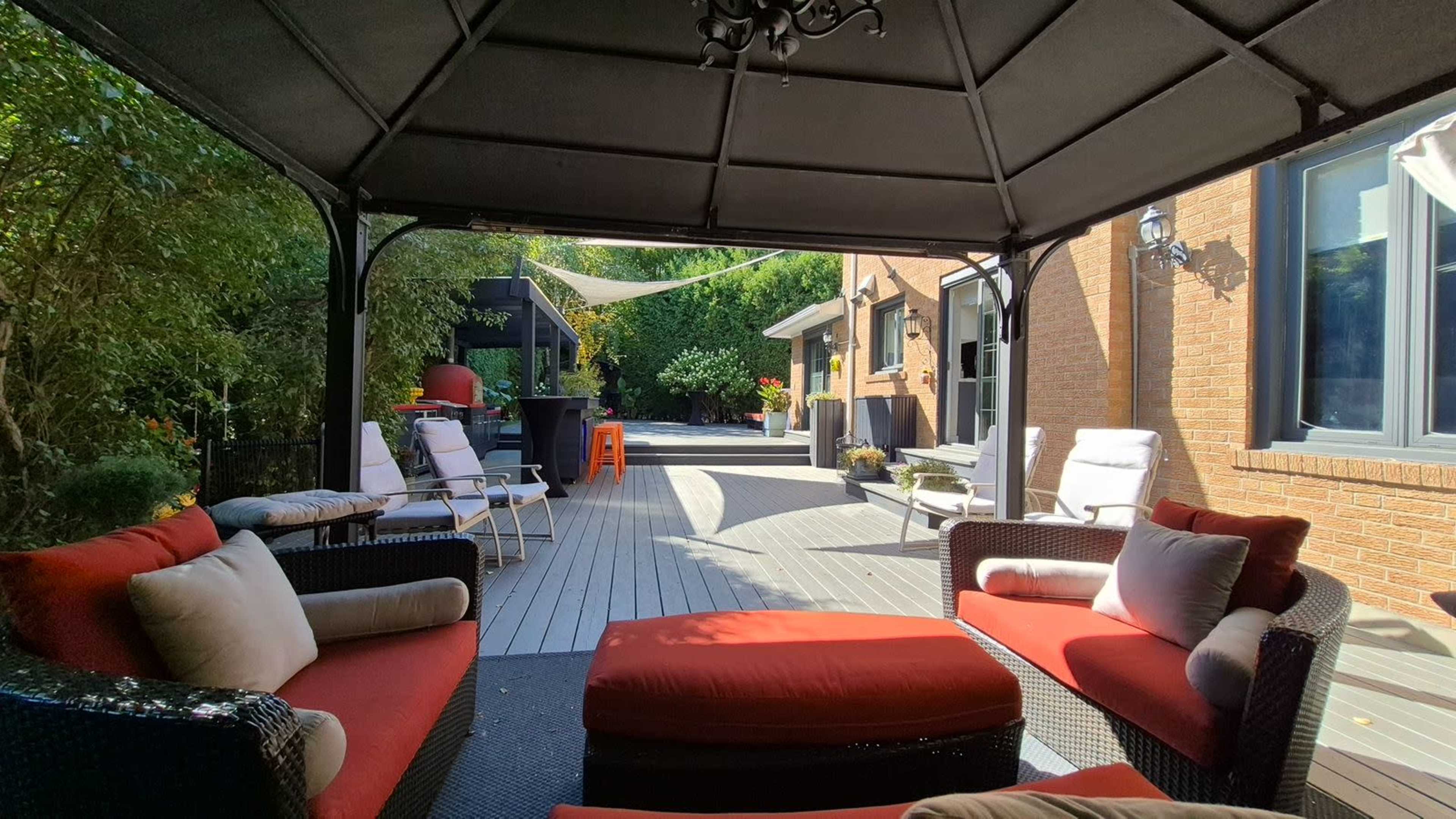 A shaded patio features red-cushioned furniture arranged around a central ottoman, leading to a wooden deck and landscaped garden area.
