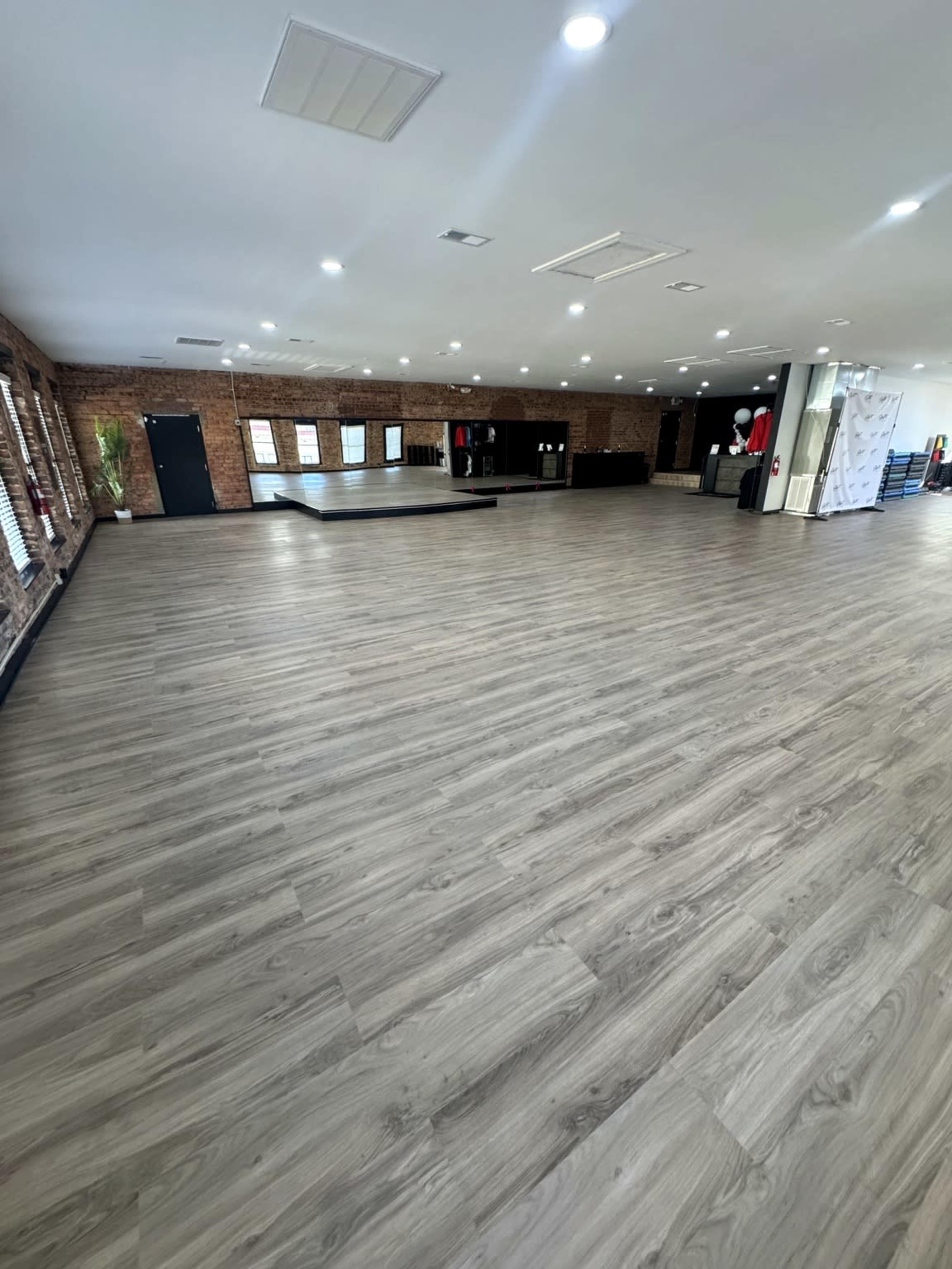 The image shows an empty, spacious interior with wooden flooring, brick walls, and soft lighting.