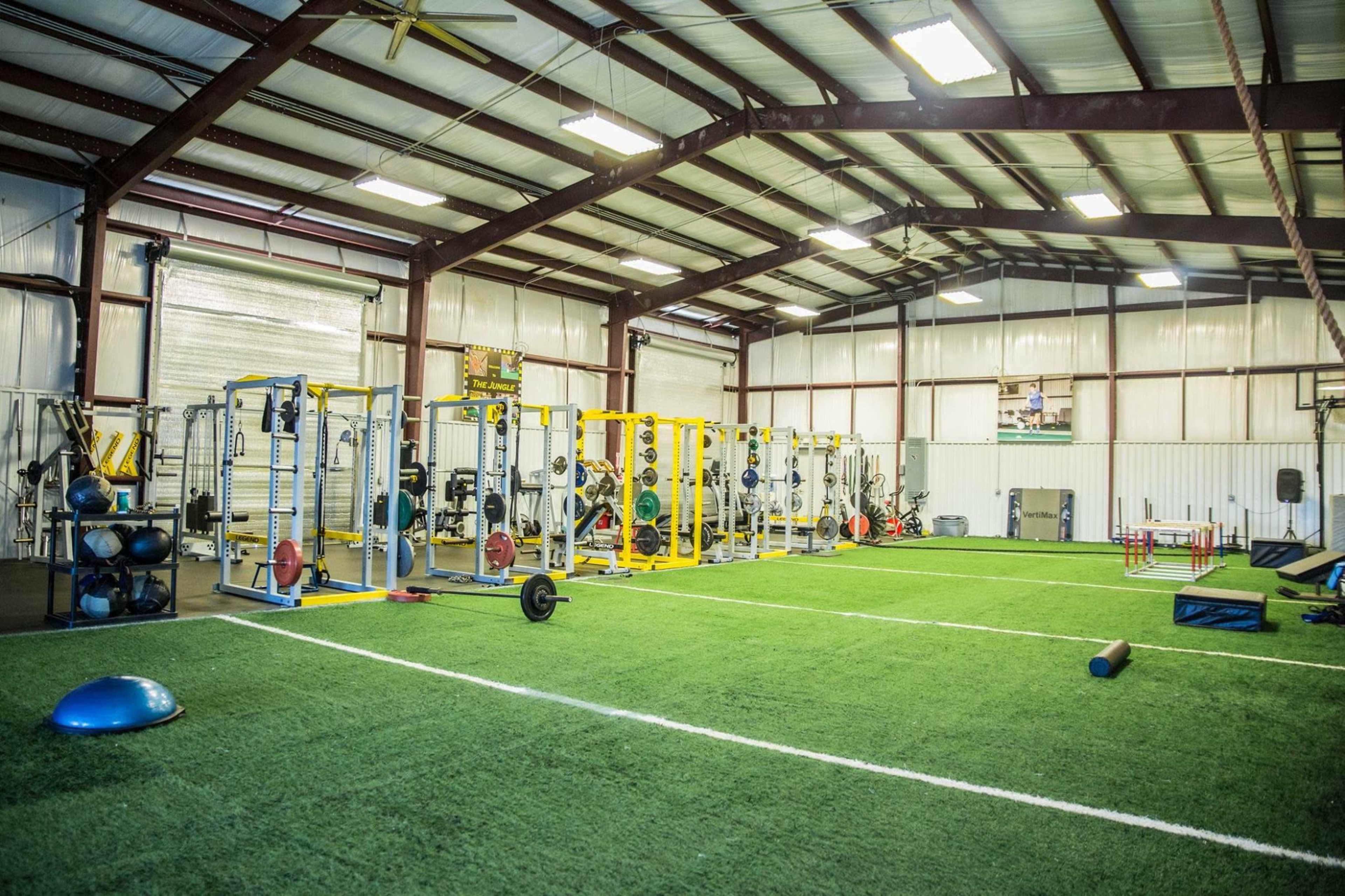 A spacious indoor gymnasium equipped with weightlifting stations, exercise equipment, and artificial turf flooring.