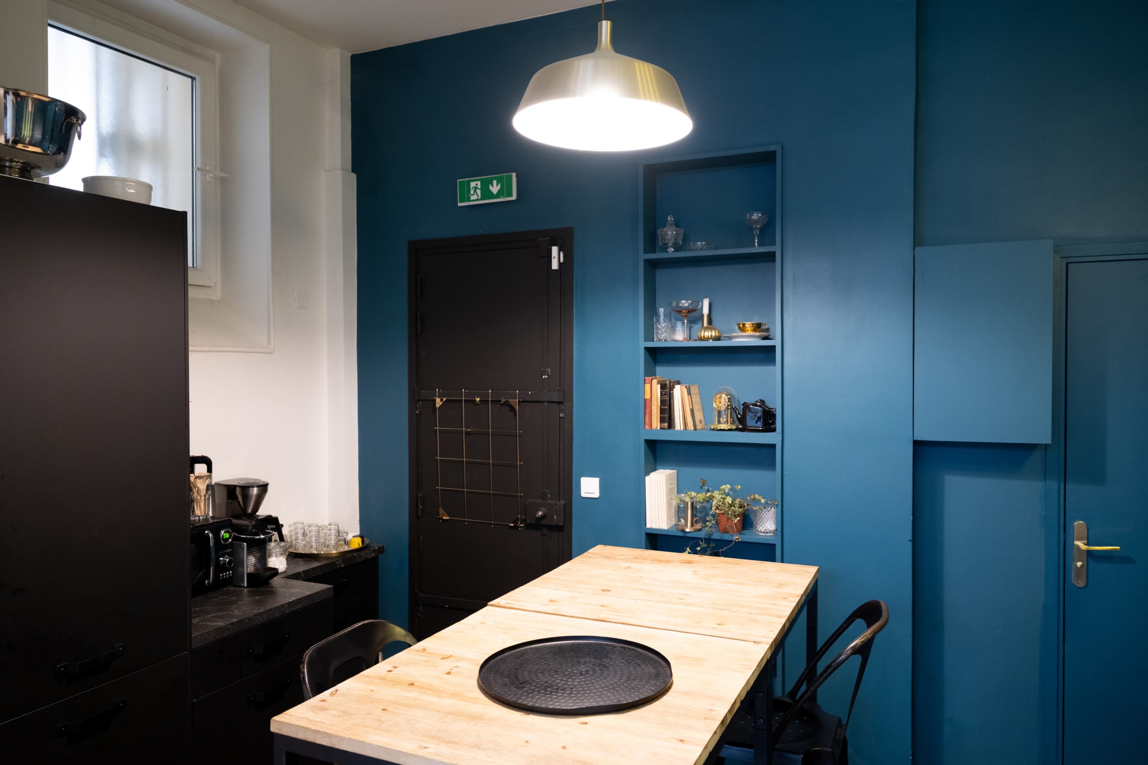 A modern kitchen features a wooden table, black cabinets, and a blue wall with shelves holding books and decorative items.