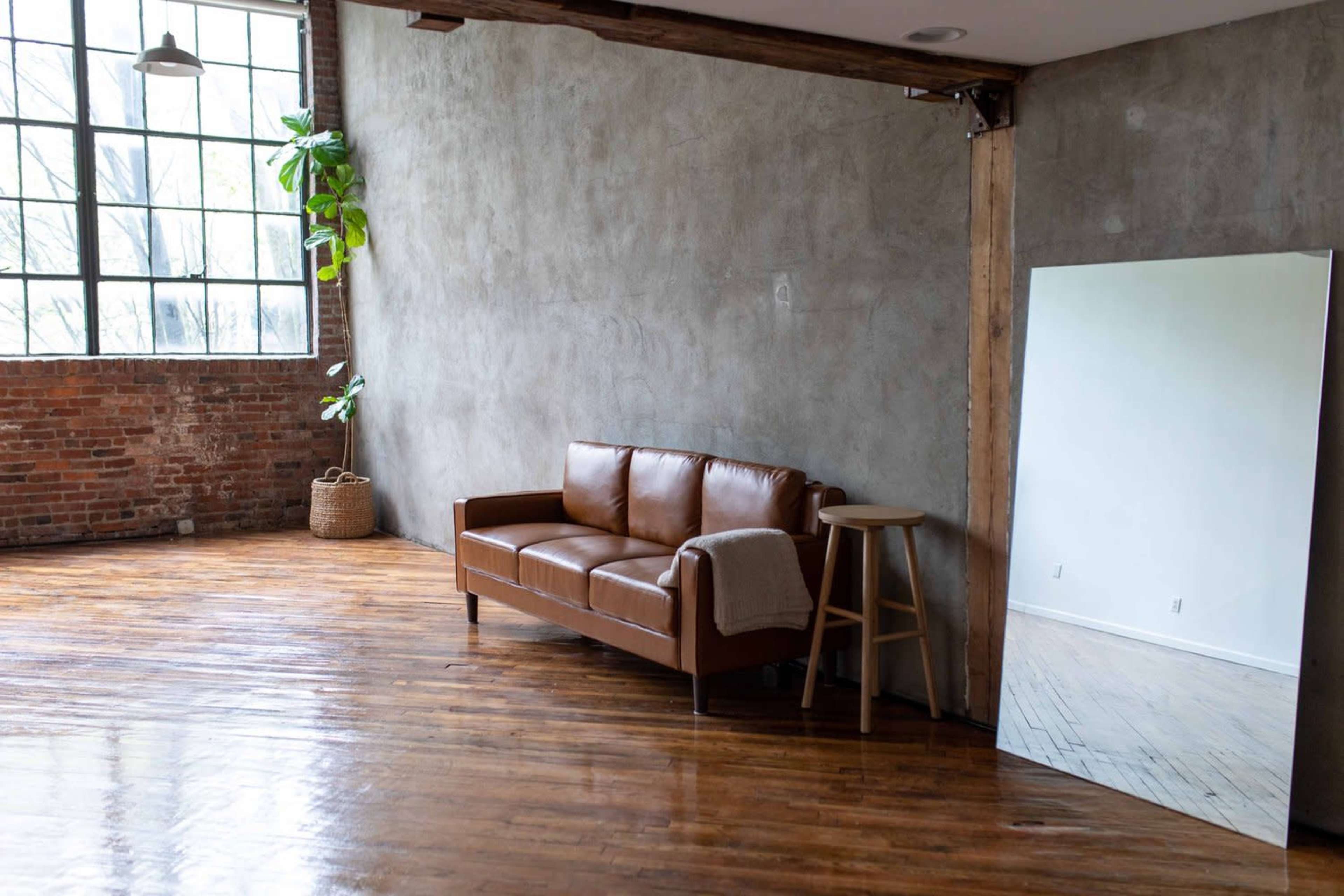 The image shows a spacious room with a brown leather sofa, a small wooden stool, a large mirror, and a concrete wall, complemented by a potted plant near a large window.