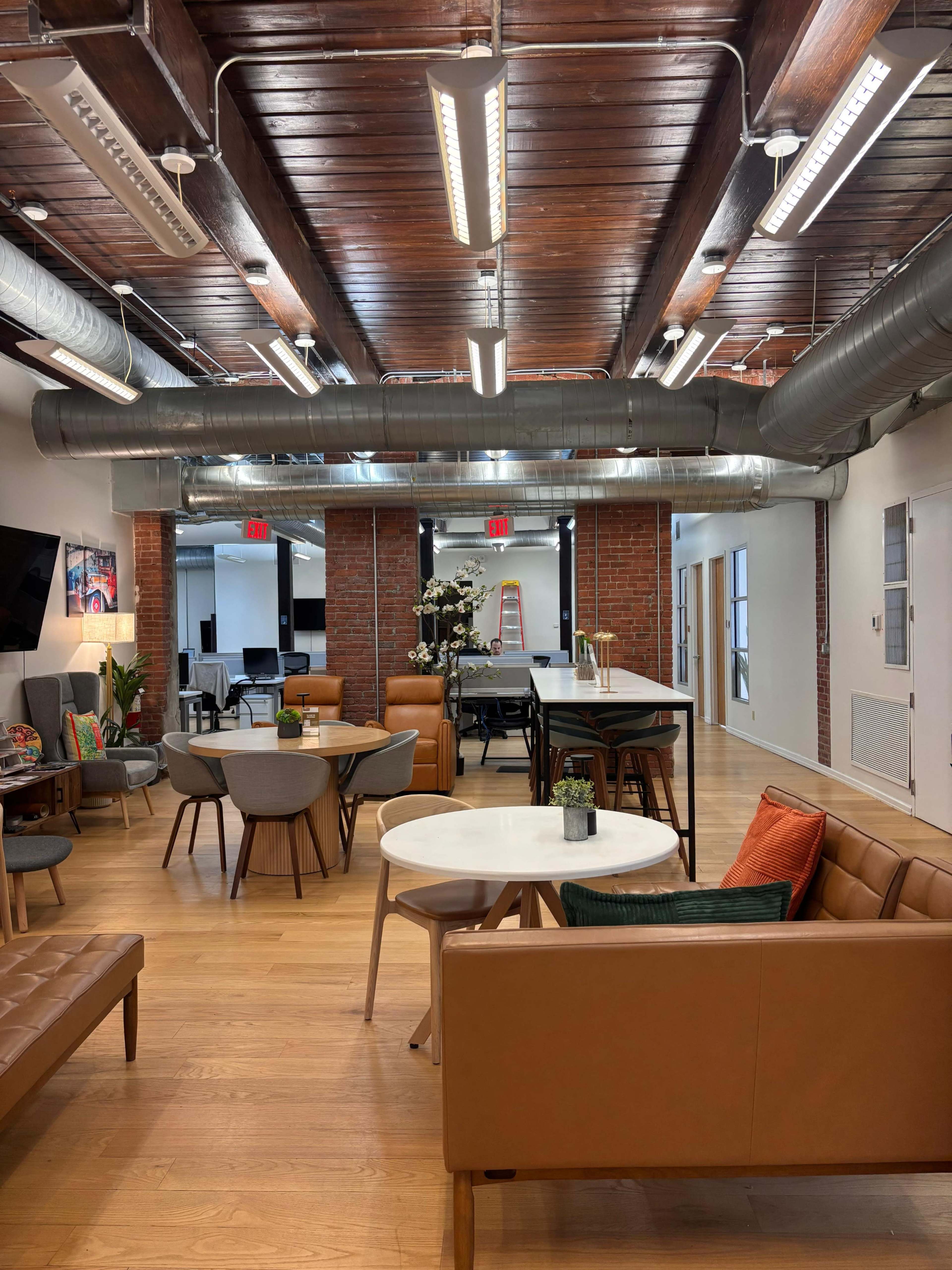 The image shows an open office space with wooden beams, a mix of seating areas, and a lounge-like atmosphere, featuring plants and modern furnishings.